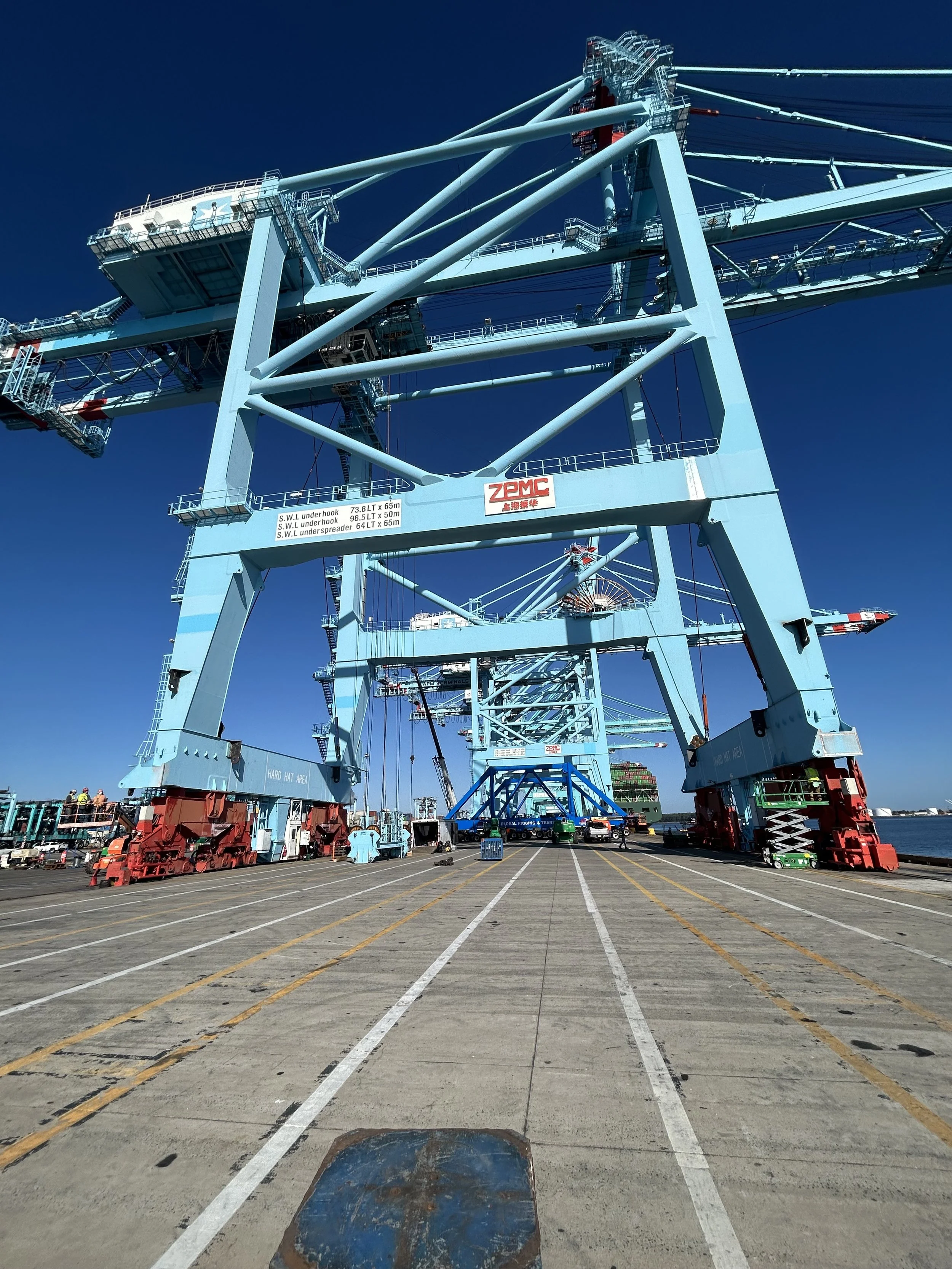 Large blue crane on a dock against a clear blue sky with some people and vehicles in the foreground.