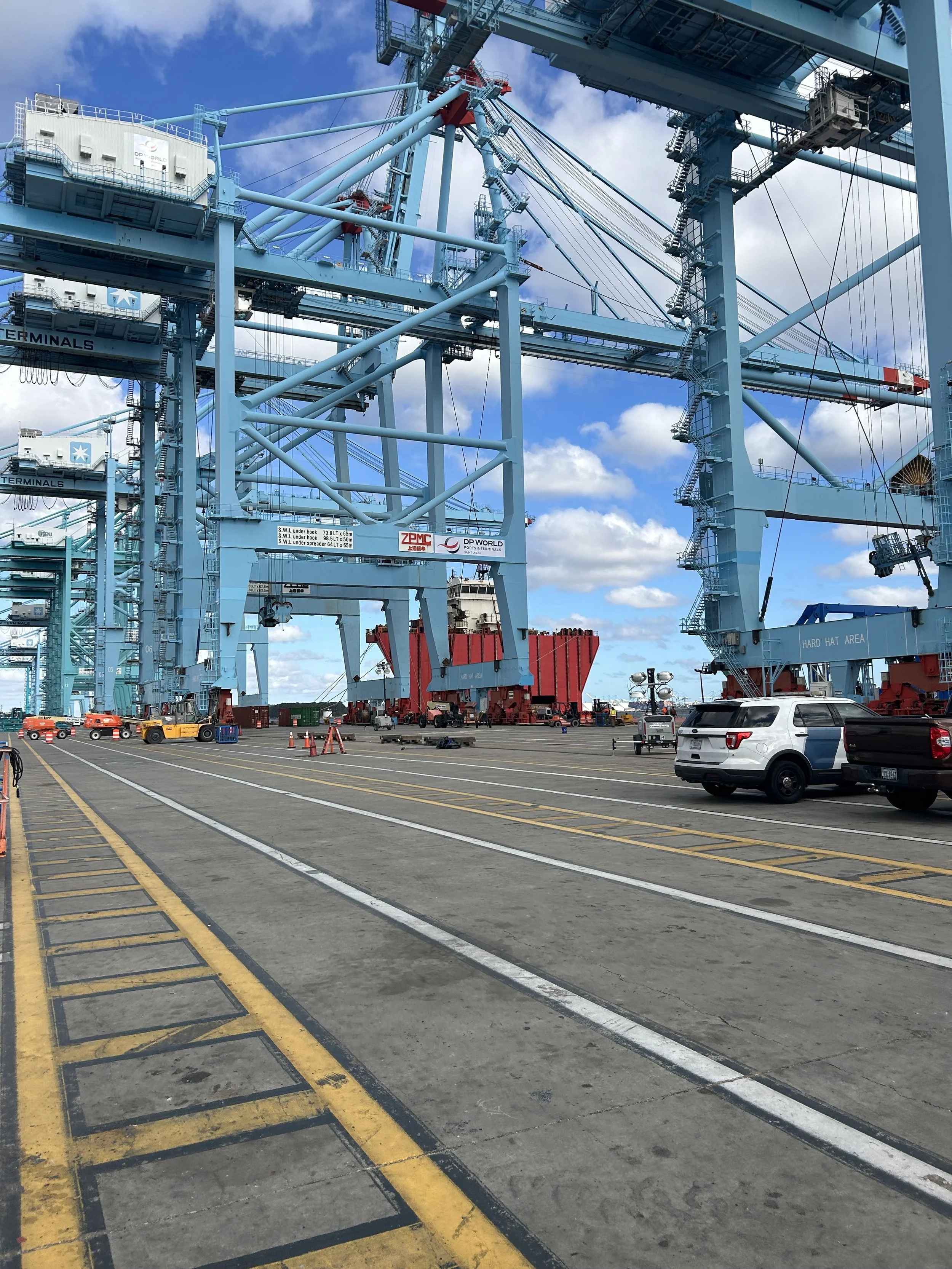 Crane at a port with shipping containers and vehicles below, blue sky with clouds.