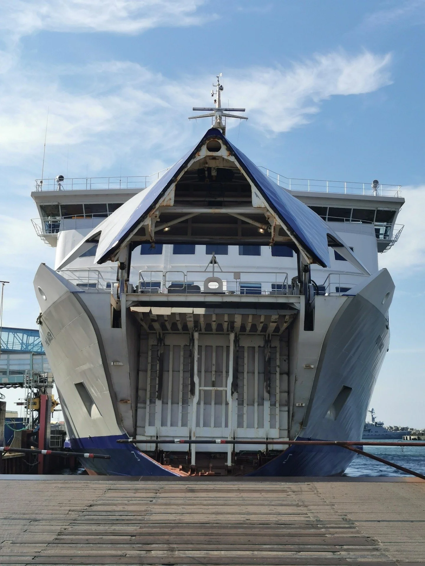View of the front of a large cruise ship docked at a port, with the bow facing the camera, under a partly cloudy sky.
