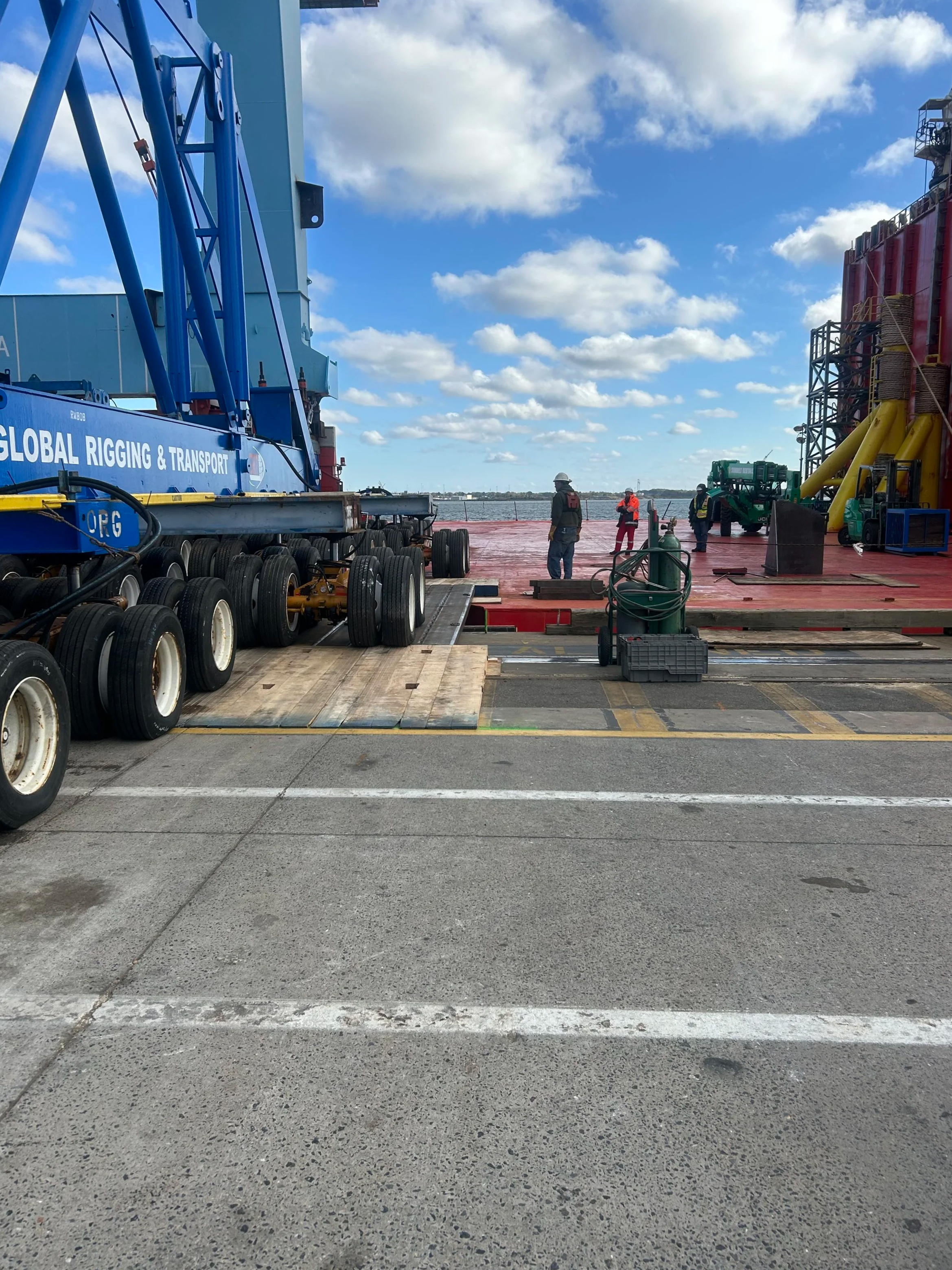 Construction workers standing on a dock next to industrial equipment, with a body of water and partly cloudy sky in the background.