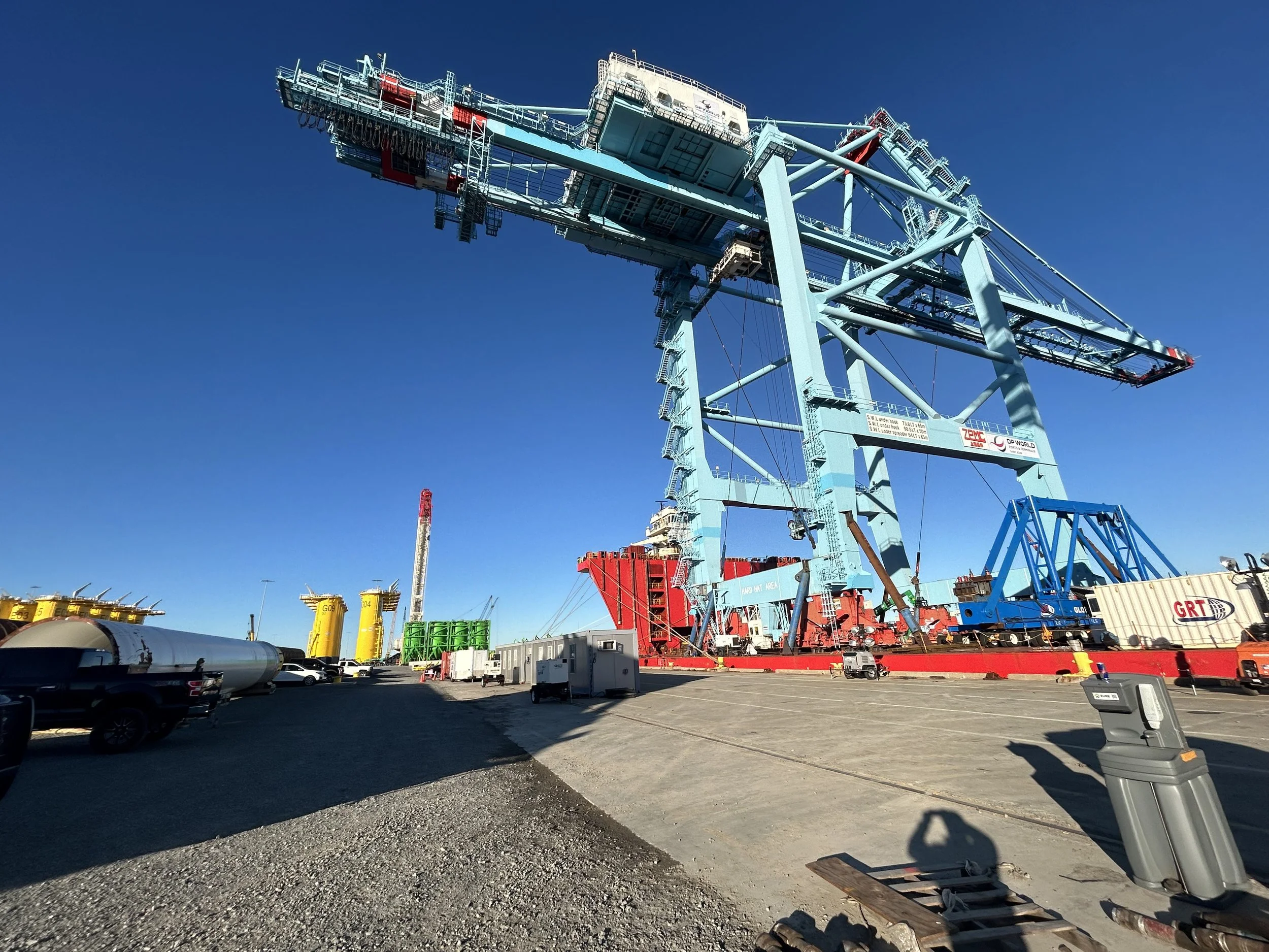 A large industrial crane at a construction or shipping site with a clear blue sky in the background. The area has parked vehicles, containers, and equipment.