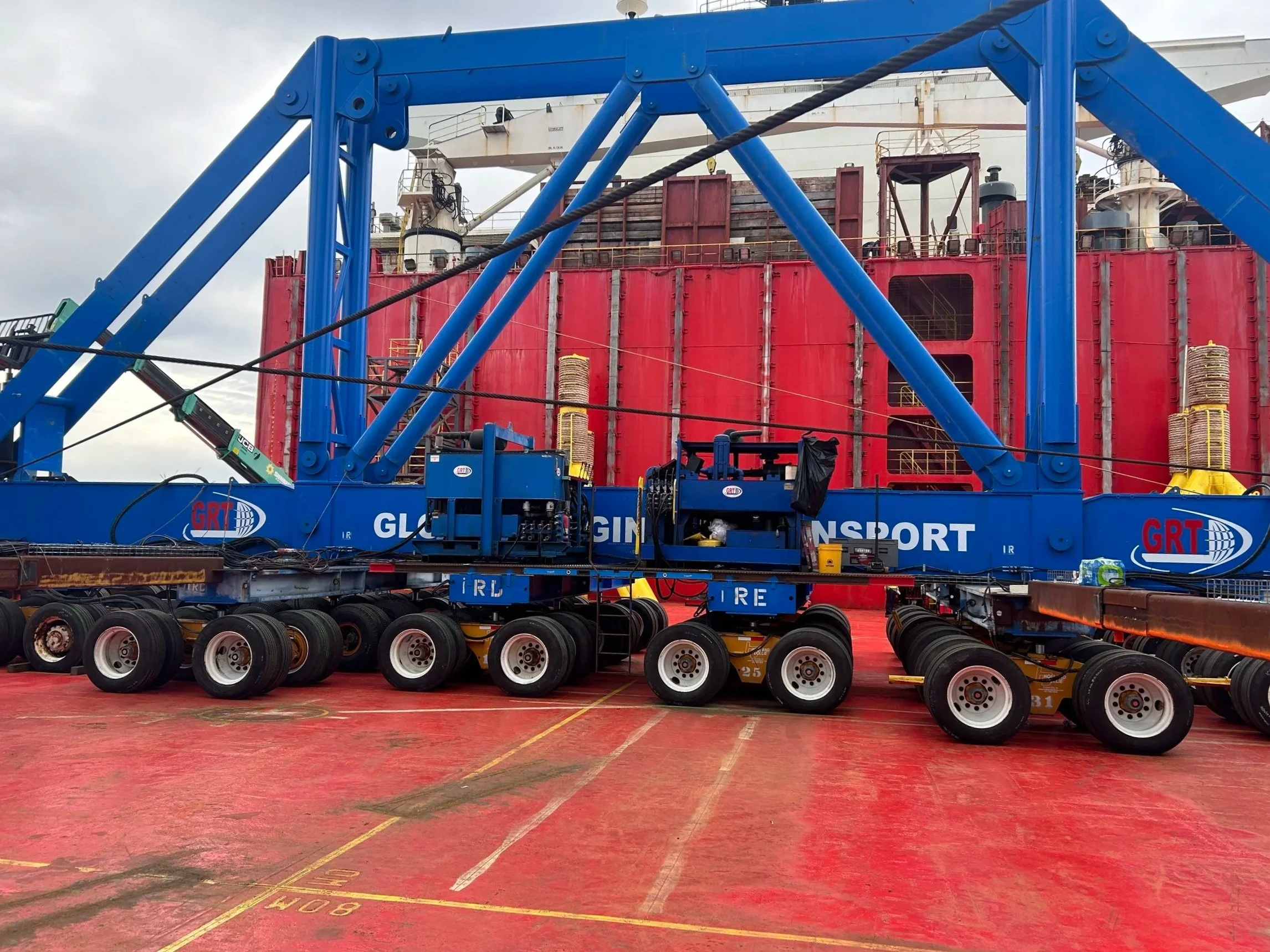 A blue industrial support structure with wheels and machinery. Behind it is a large red ship with various equipment and railings on its deck. The scene appears to be at a port or dock, with a cloudy sky overhead.