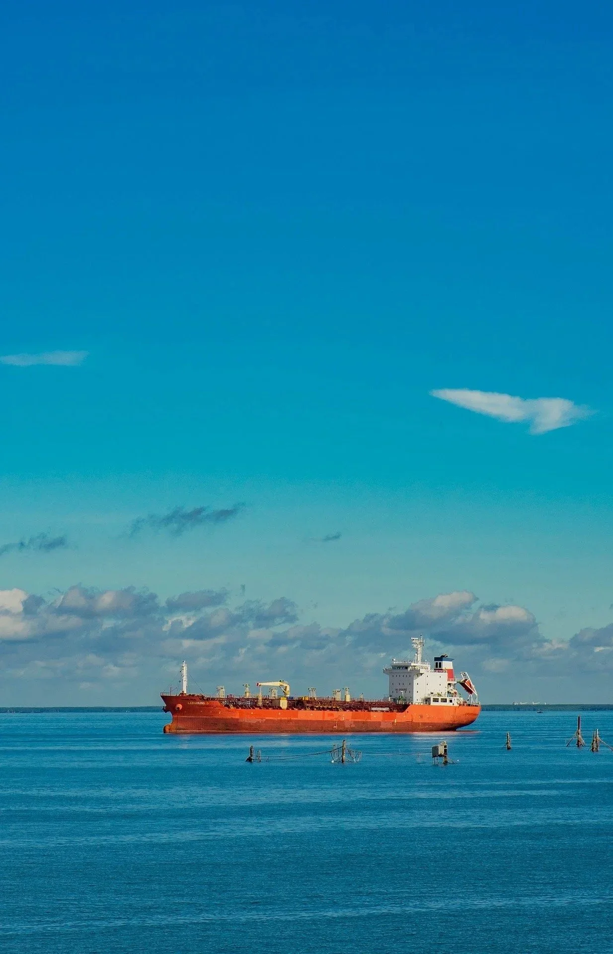 A large red cargo ship floating on calm blue ocean water under partly cloudy sky.