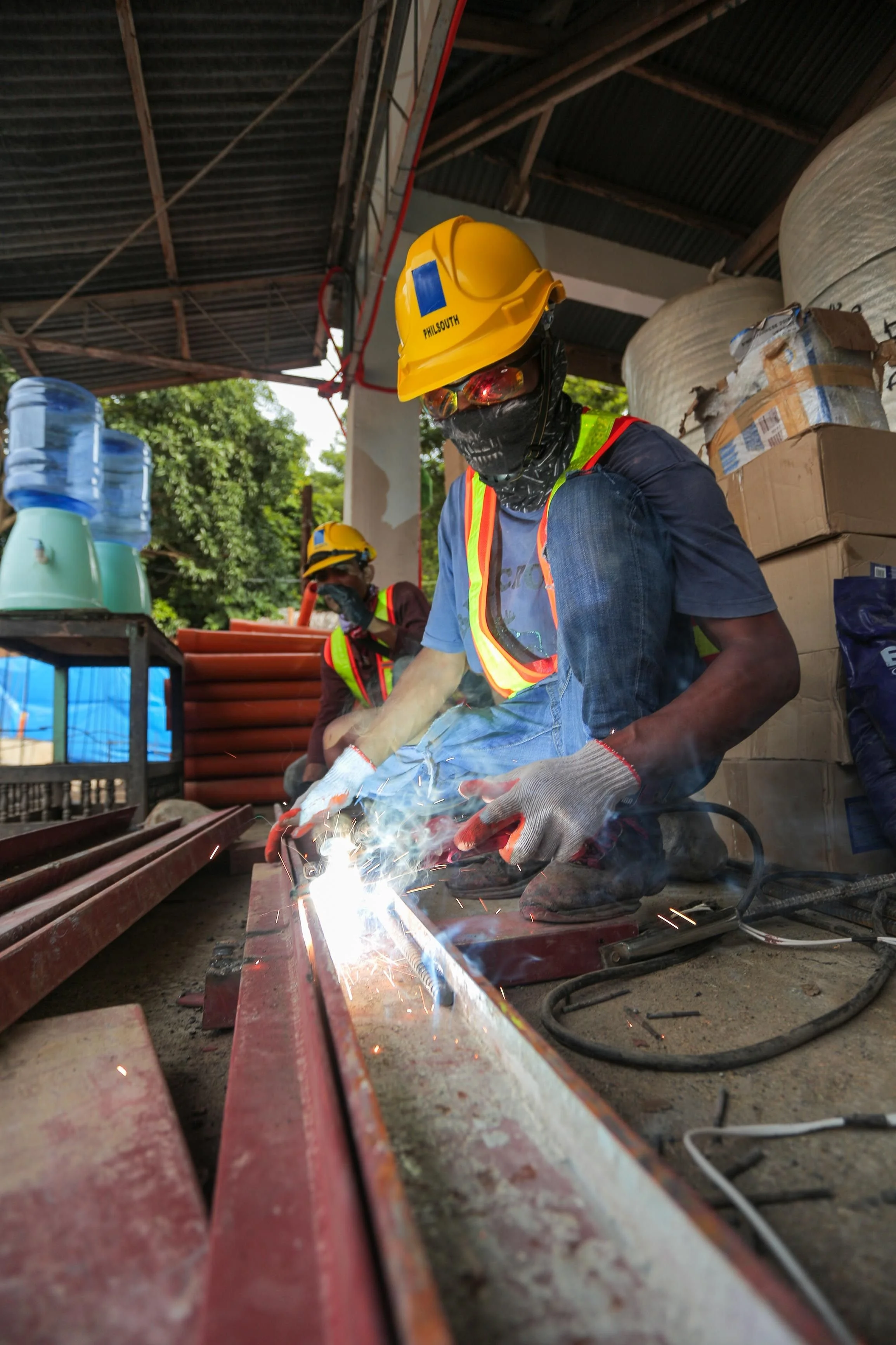 Worker welding metal pipe, wearing yellow helmet, goggles, gloves, and safety vest, with another worker in the background, in an industrial workshop with boxes and water bottles.