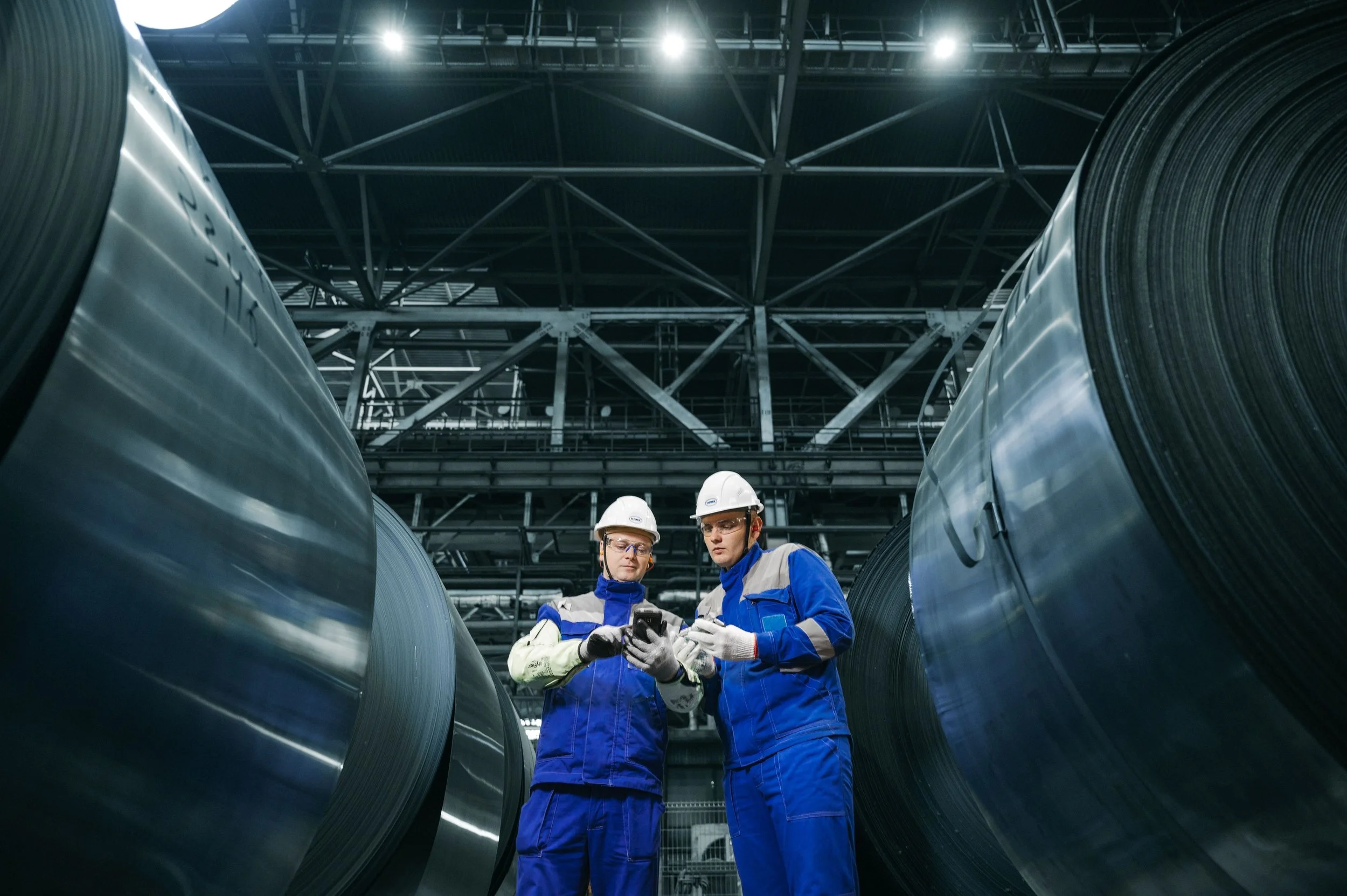 Two workers in blue uniforms and white helmets standing between large metal rolls in an industrial warehouse, inspecting a device or document.