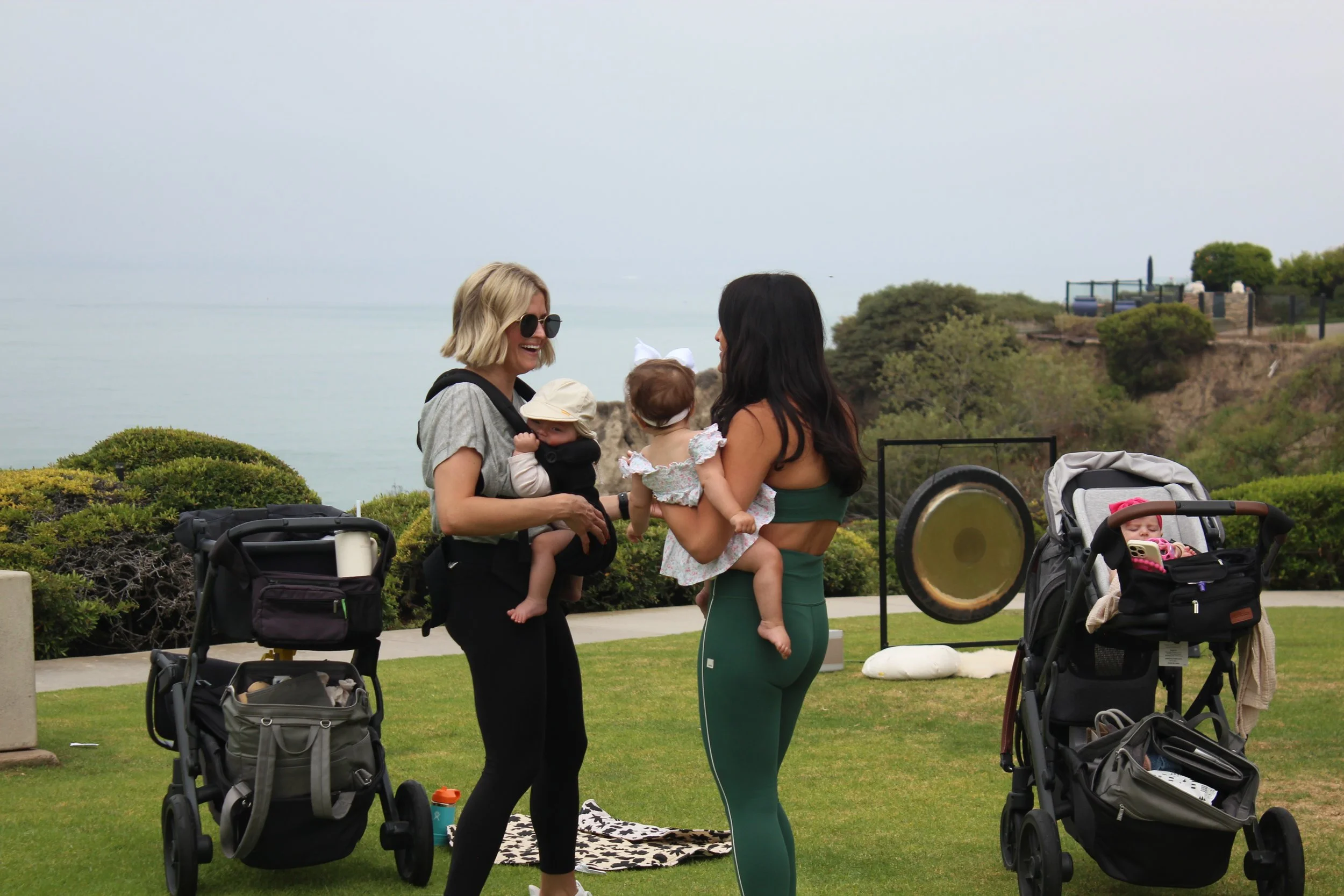 Two women holding babies and after using their stroller as their barre and working out with The Stroller Edit in Orange County California near strollers and a large drum, with a scenic Pacific ocean and greenery in the background.