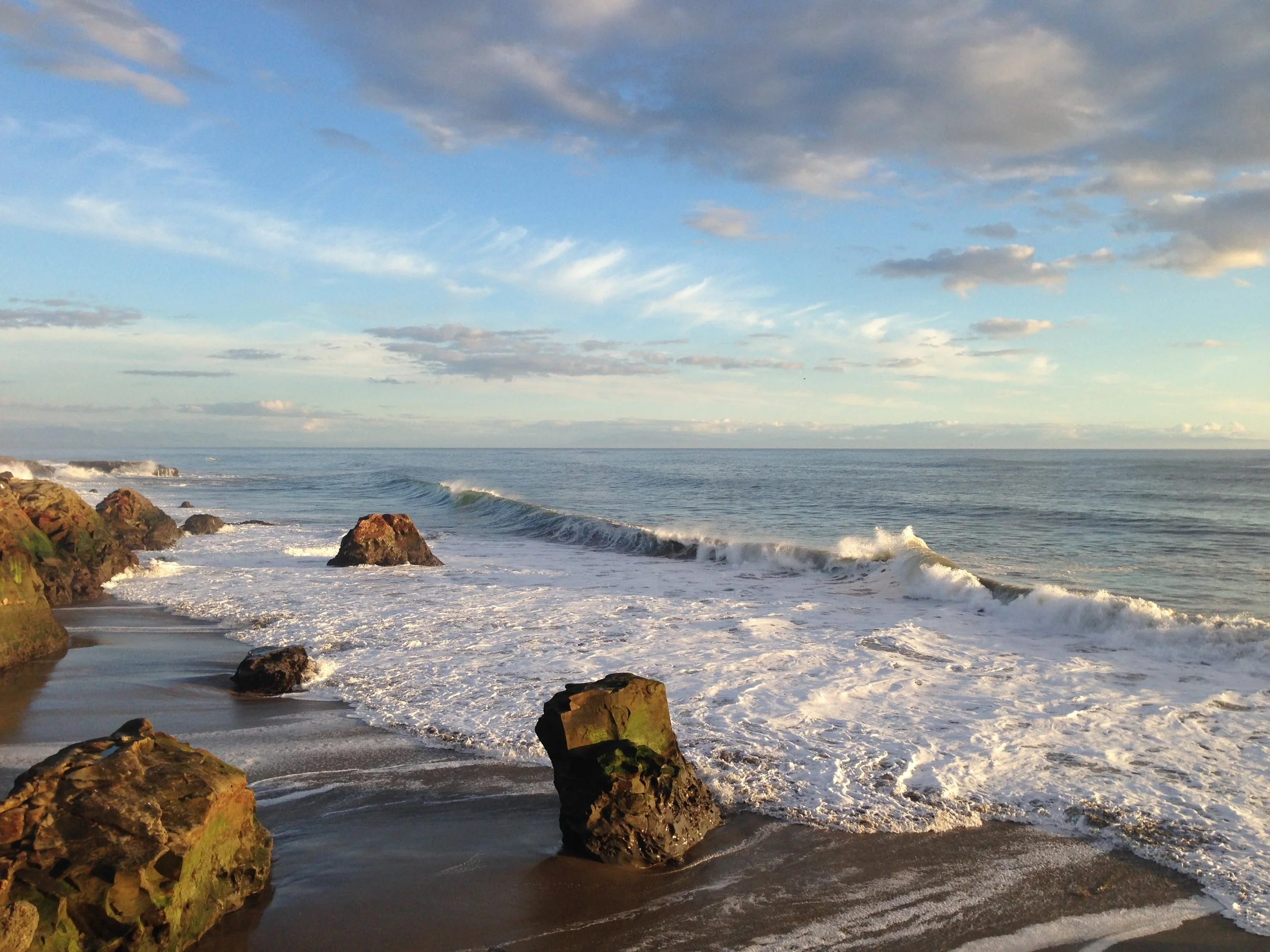 A scenic view of a beach with rocks in the foreground, gentle waves and an expansive sky with scattered clouds in the background. This coastal region is where Santa Cruz Garlic grows locally adapted seed garlic.