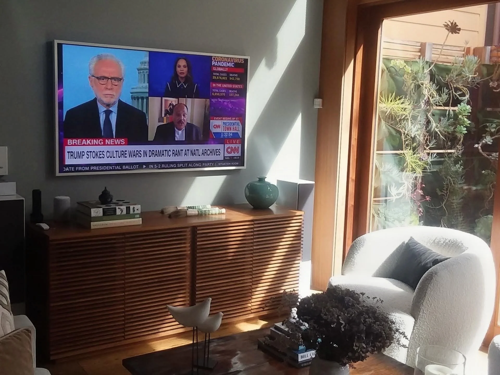 Living room with a wall-mounted flat screen TV showing a news broadcast, a white armchair with a dark pillow, a wooden sideboard, a green ceramic vase, and a window with sunlight and plant view.