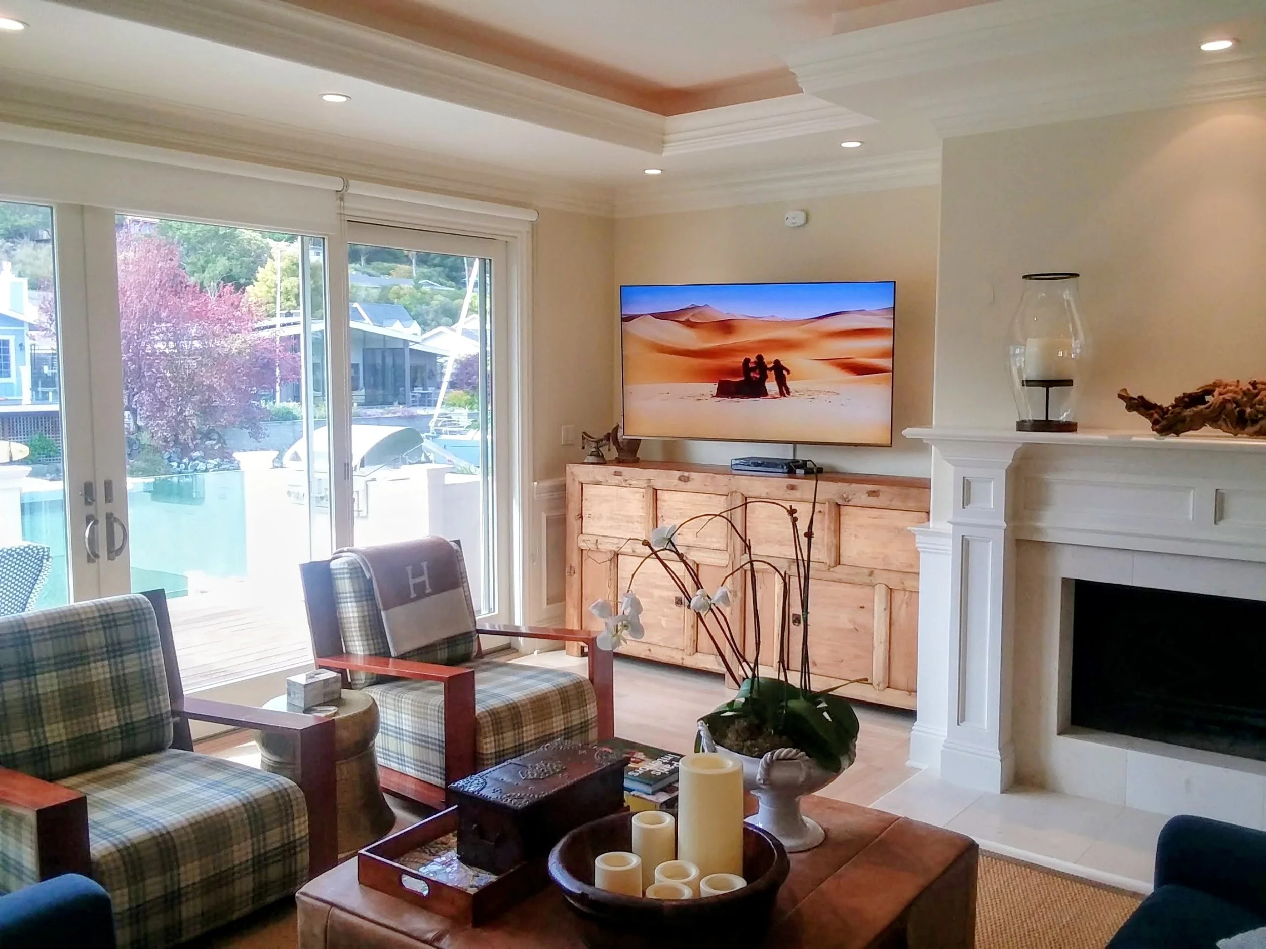 Living room with plaid armchairs, a wooden coffee table with candles, a TV showing desert landscape, large sliding glass doors with trees outside, and a white fireplace with decorative items.