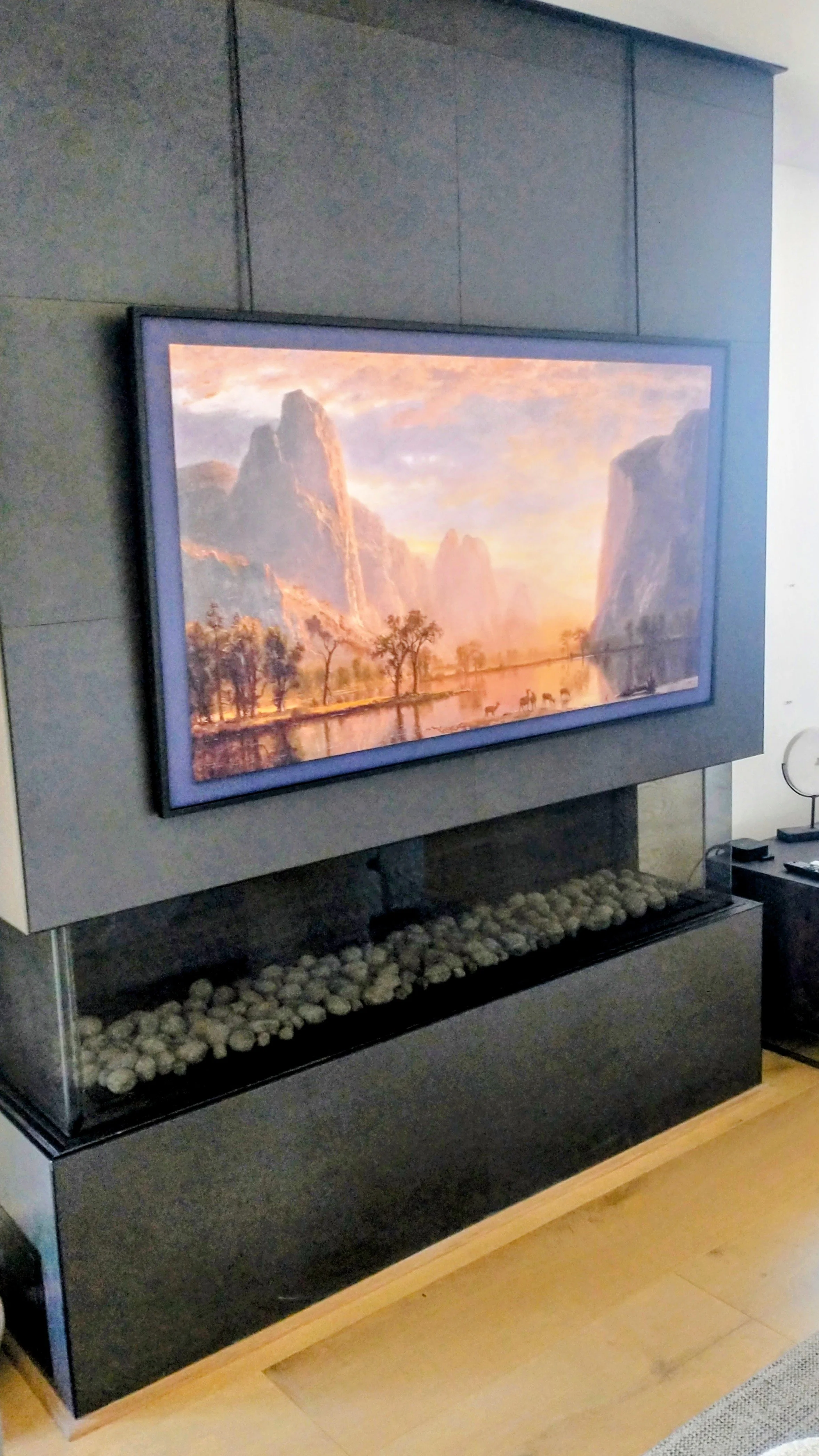 A framed landscape painting hanging on a gray wall above a modern fireplace filled with white rocks.
