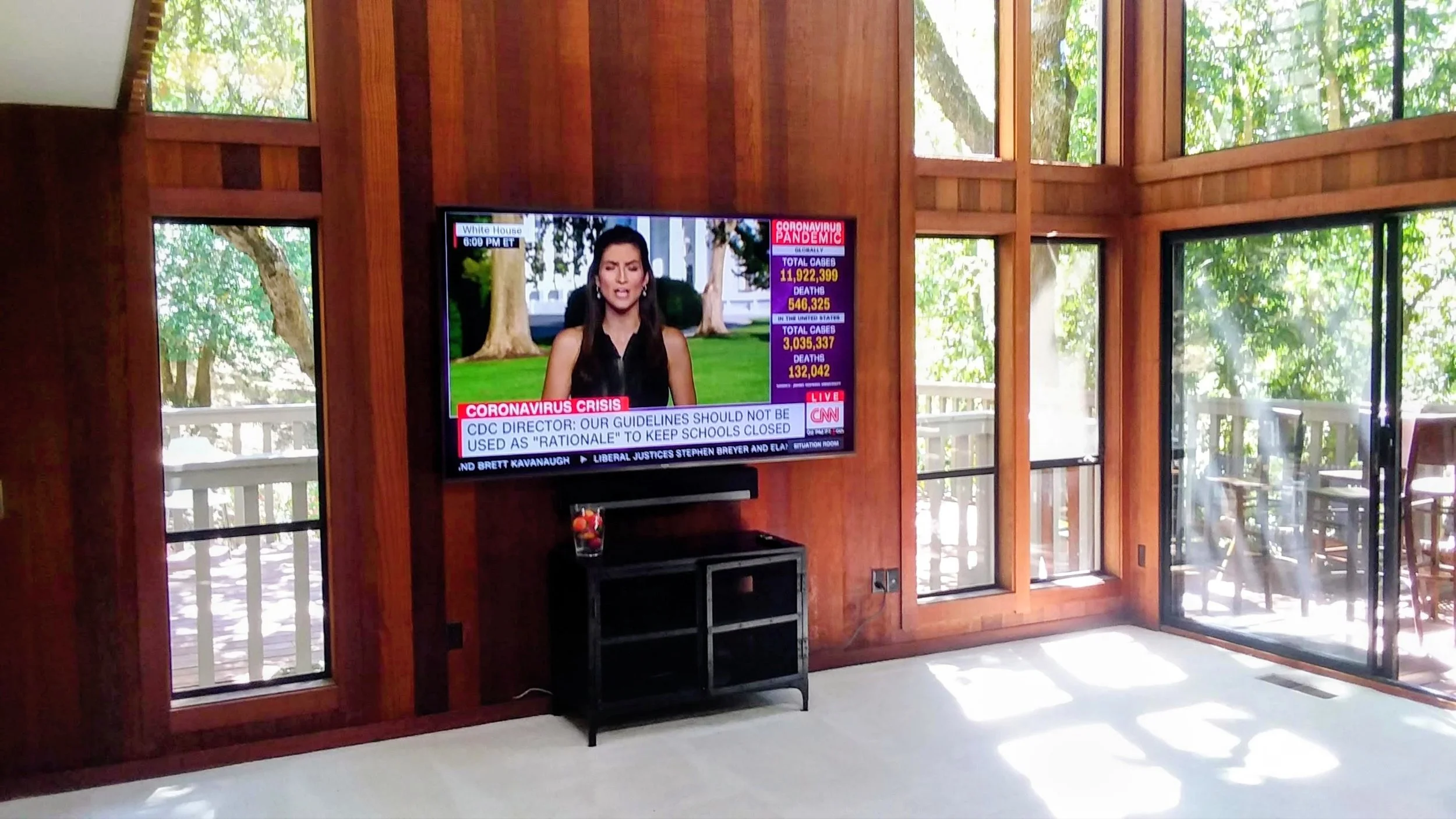 A flat-screen TV mounted on a wooden wall, displaying a news broadcast about the COVID-19 pandemic, with text and graphics on the right side of the screen. The room has large glass windows and doors with views of trees outside, and a small black cabinet underneath the TV.
