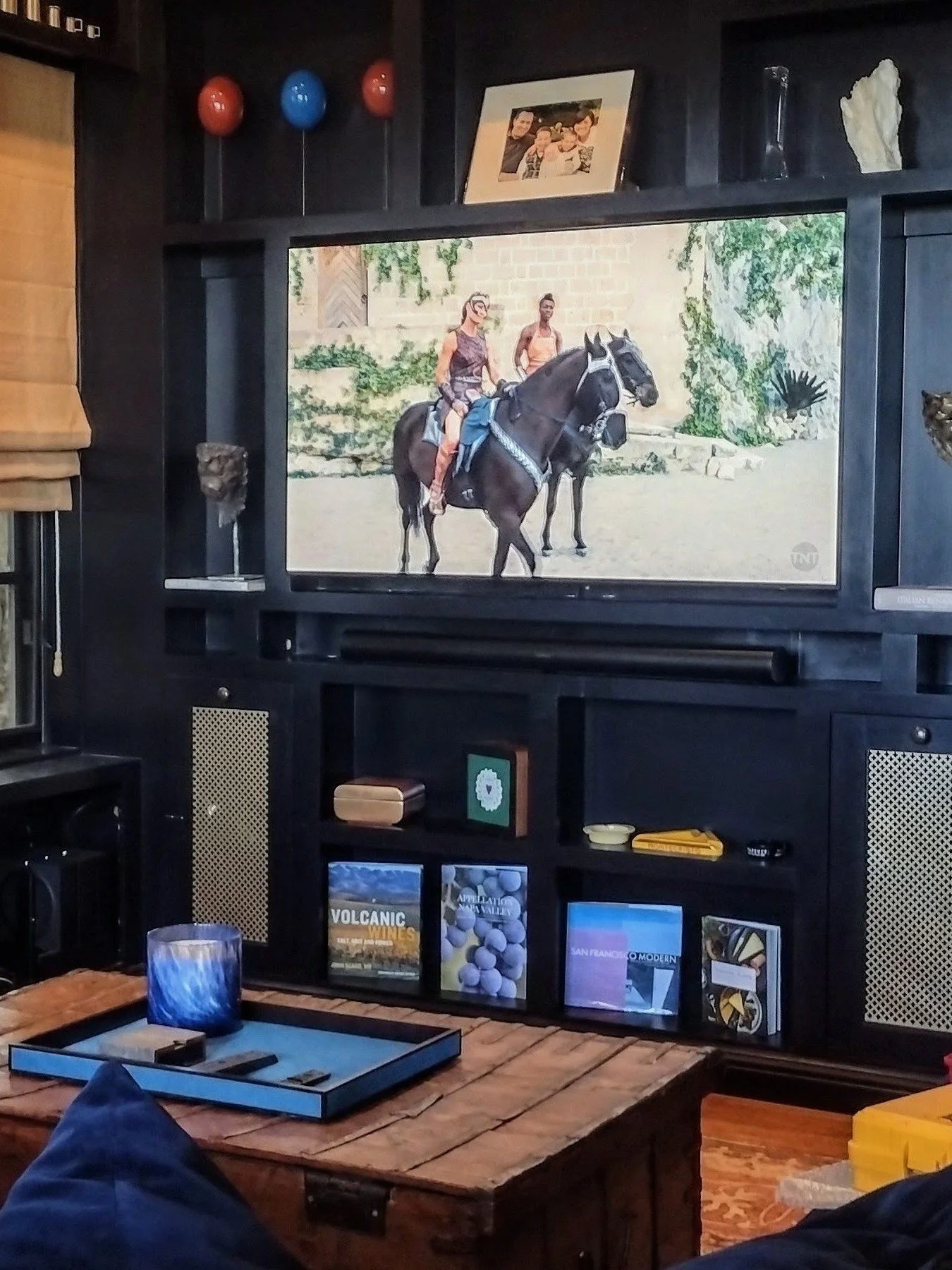 Living room with a black entertainment center holding books, magazines, and decorative items. On the wall, there's a large TV displaying two people riding horses in an outdoor setting. Above the TV, a picture of a family is on the shelf, and there are decorative objects, including balloons and a sculpture, on the shelves