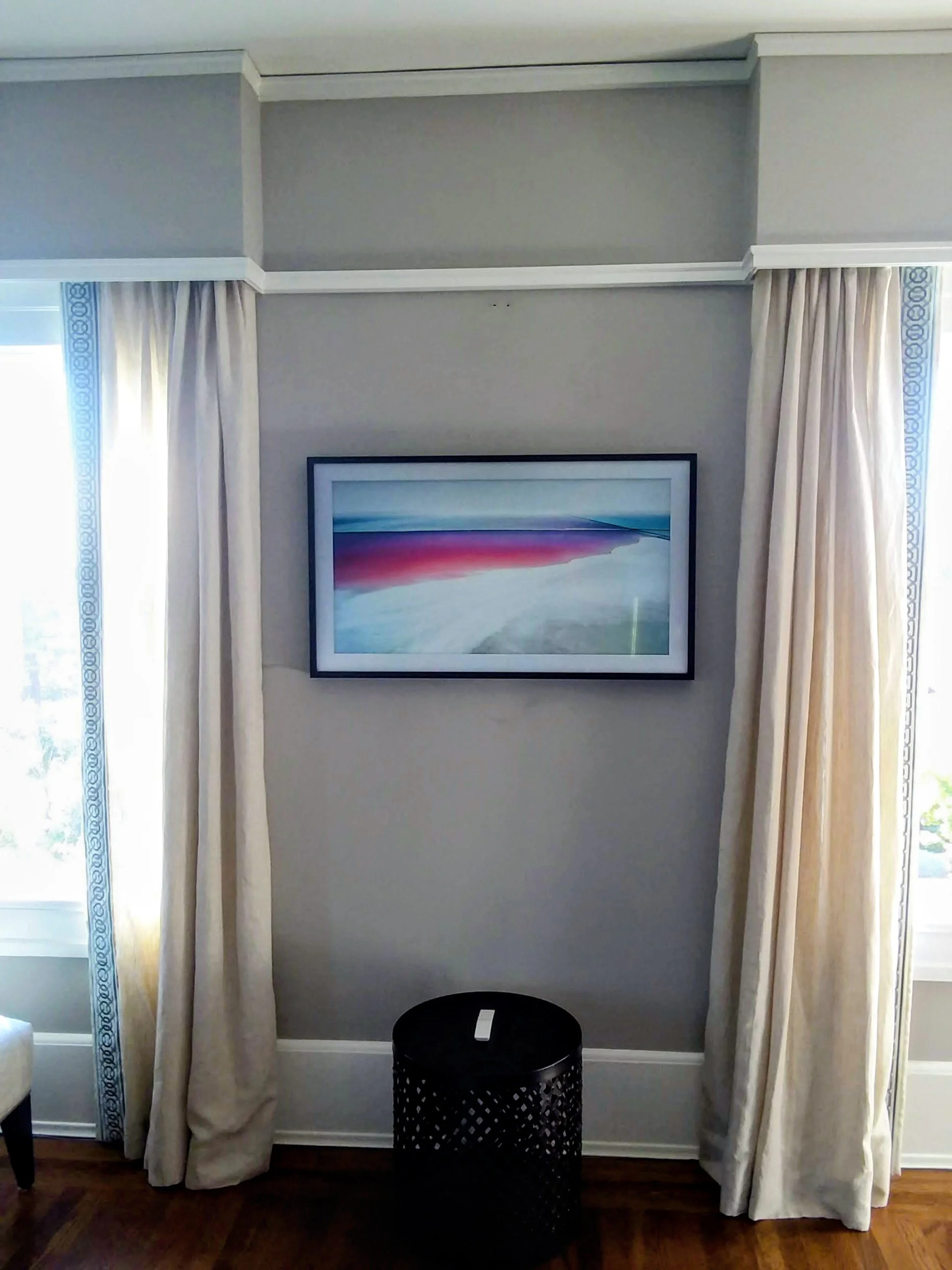 A framed abstract painting hanging on a gray wall, with floor-length cream curtains on either side. A black round stool is positioned in front of the wall, and the floor is hardwood.