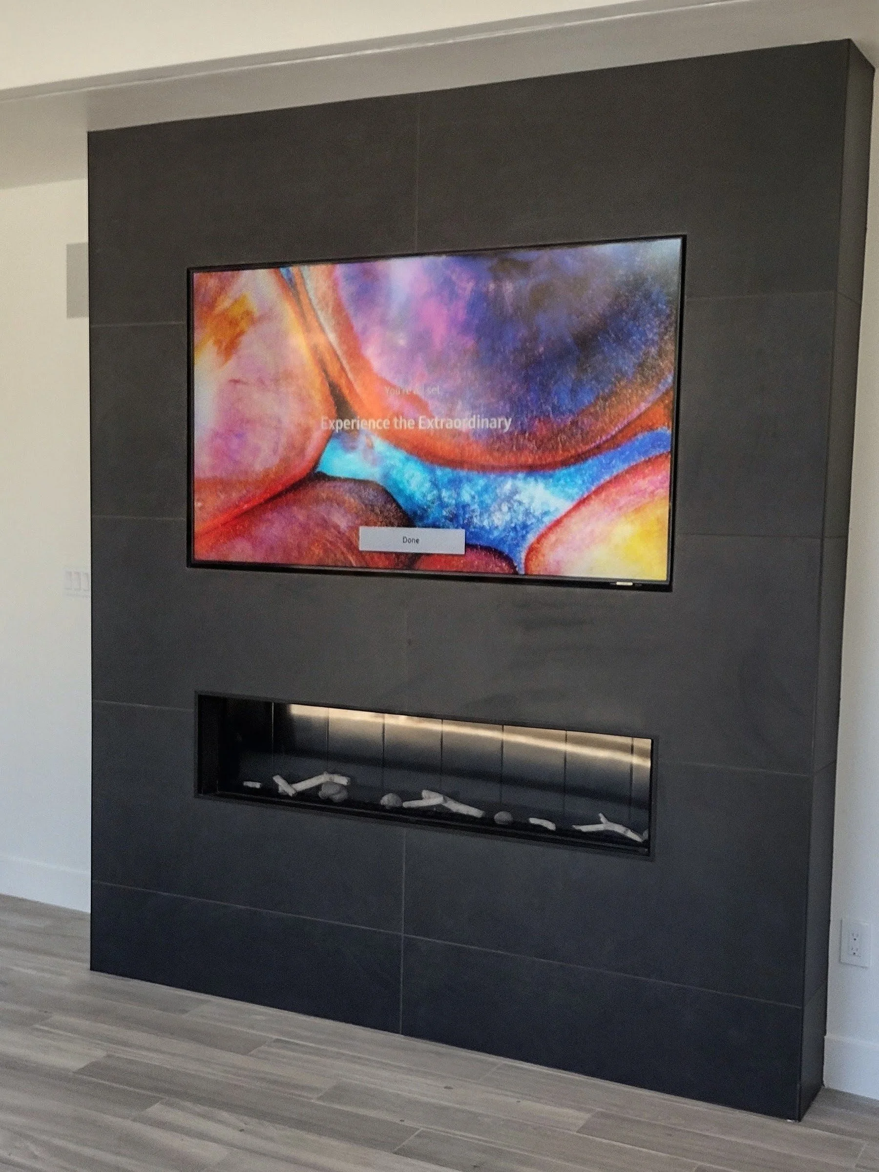 A wall-mounted flat-screen television set in a black frame on a dark gray tiled wall, with a fireplace beneath it, in a modern interior.