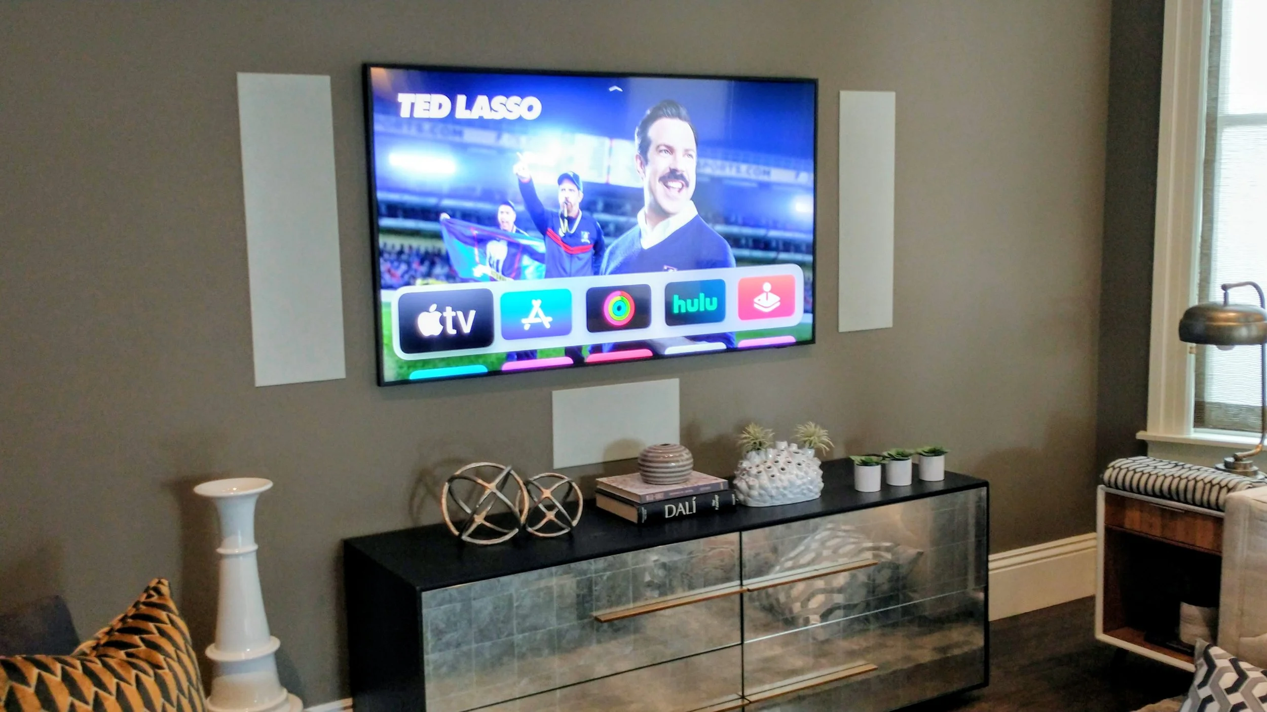 Living room with wall-mounted TV displaying a sports scene with people holding a flag, some icons like Apple TV, Hulu on the screen, and decorative items on a black and green marble console table.