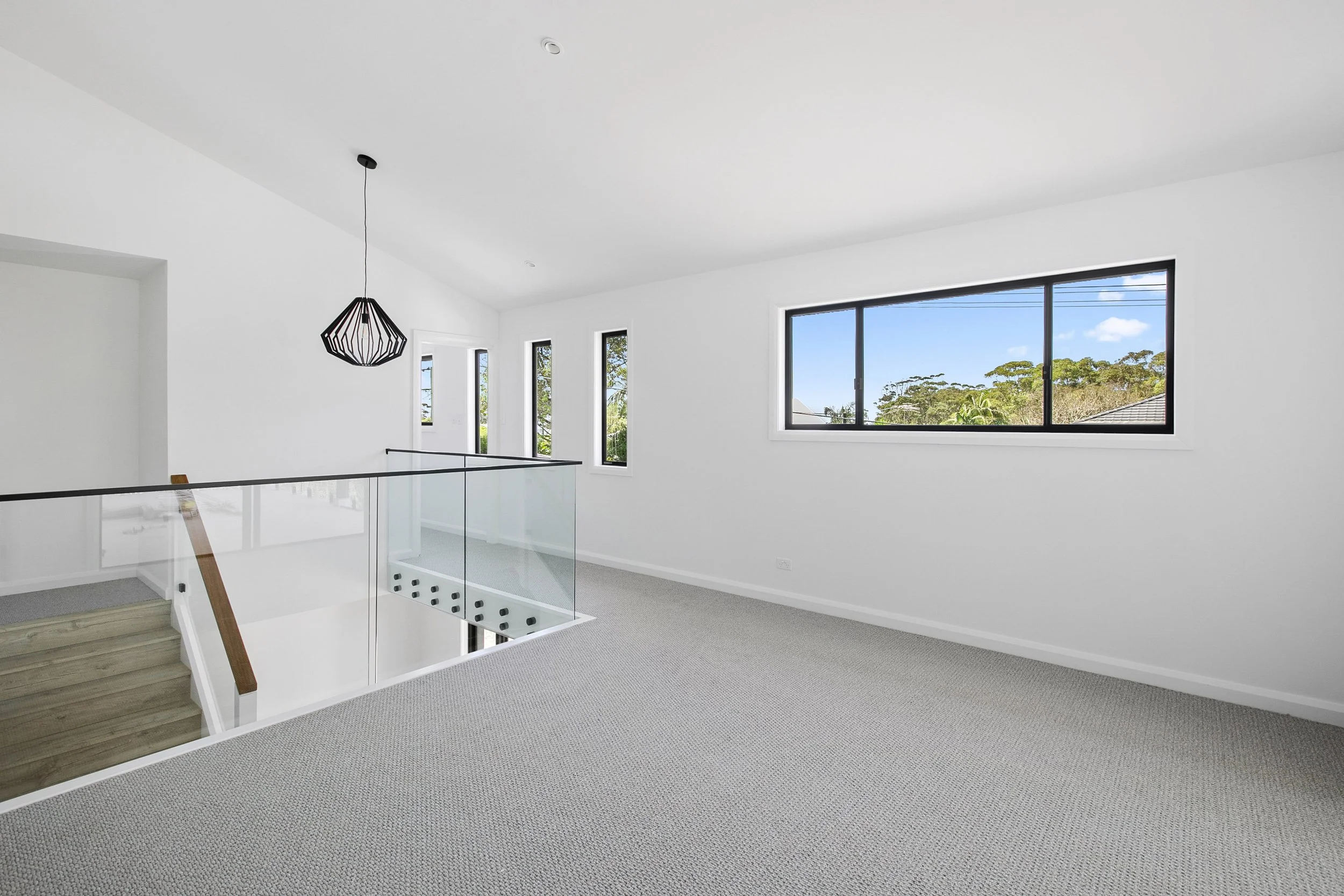 Interior view of a modern, bright room with white walls and ceiling, large windows showing a blue sky and green treetops, a sleek black pendant light, and a staircase with glass balustrade and wooden handrail.
