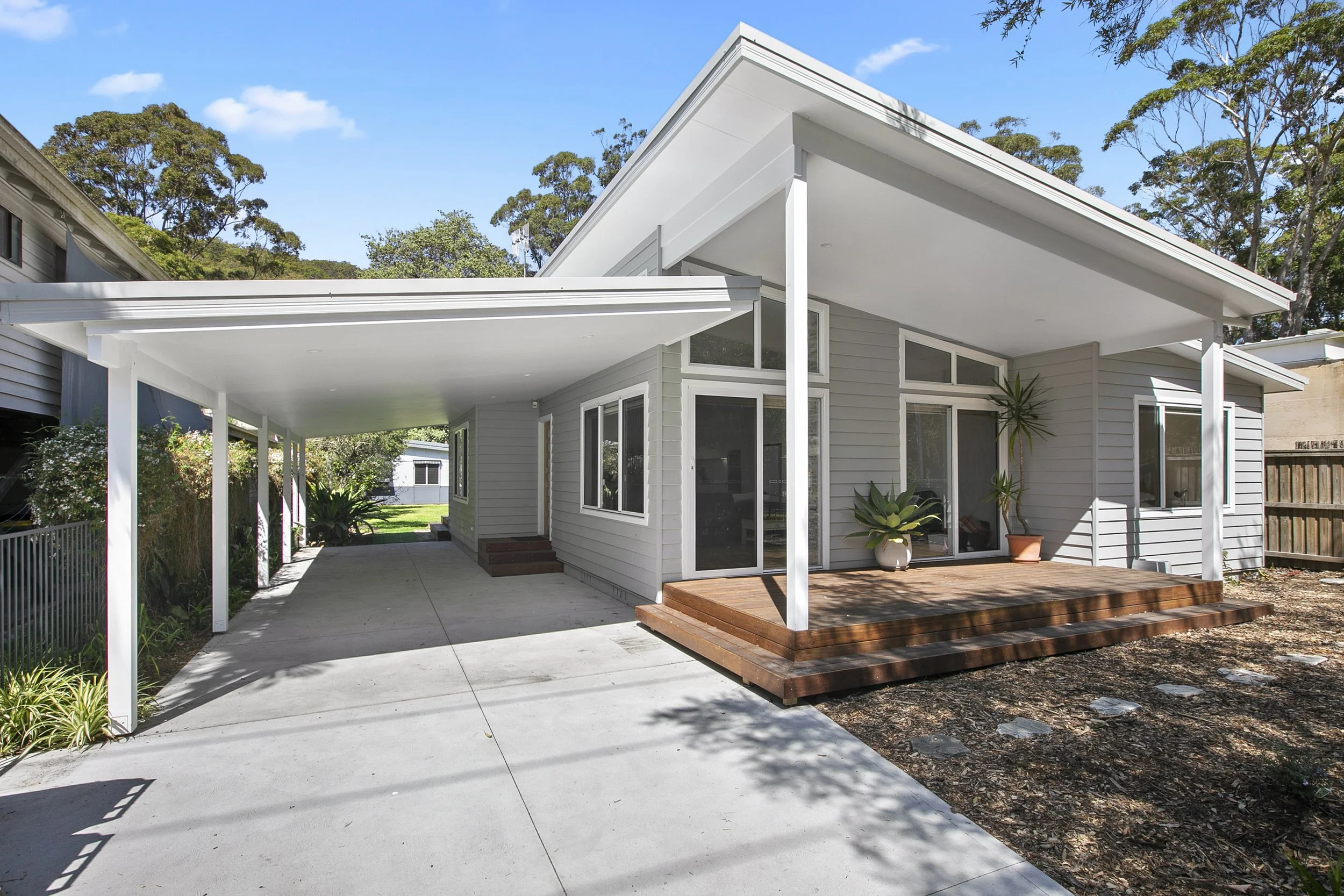 Modern house with white exterior, large windows, and a patio with a wooden deck, surrounded by green plants and trees under a blue sky.