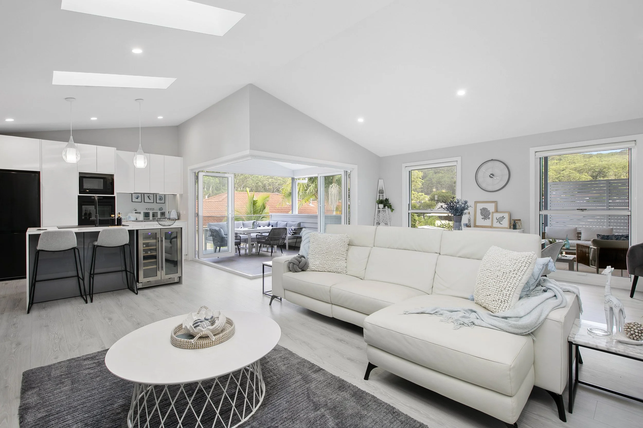 Bright, modern living room with white sofa, coffee table on dark rug, and large windows with views of greenery and neighboring houses.