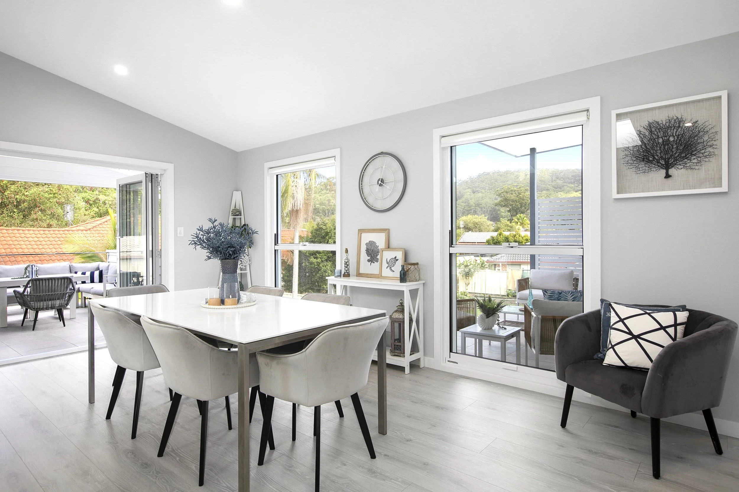 Modern living room with large windows, light-colored flooring, white walls, dining table with chairs, decorative artwork, and outdoor patio with seating.