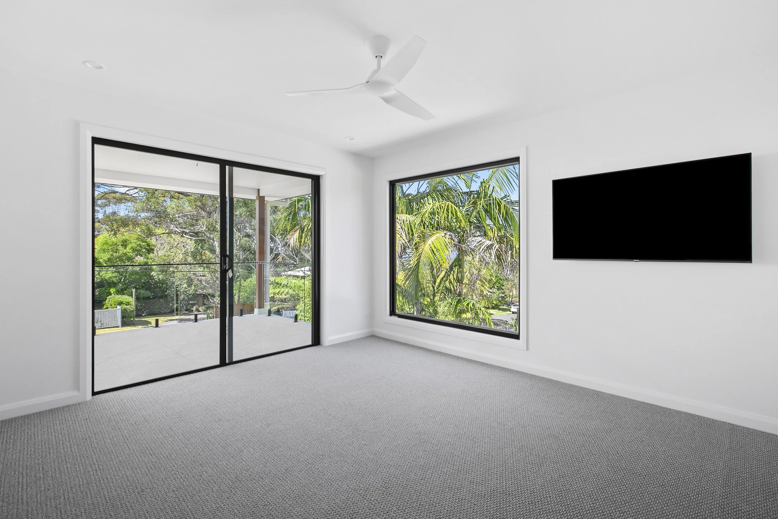 Modern living room with large windows showing trees outside, a ceiling fan, a mounted flat-screen TV, and grey carpet flooring.