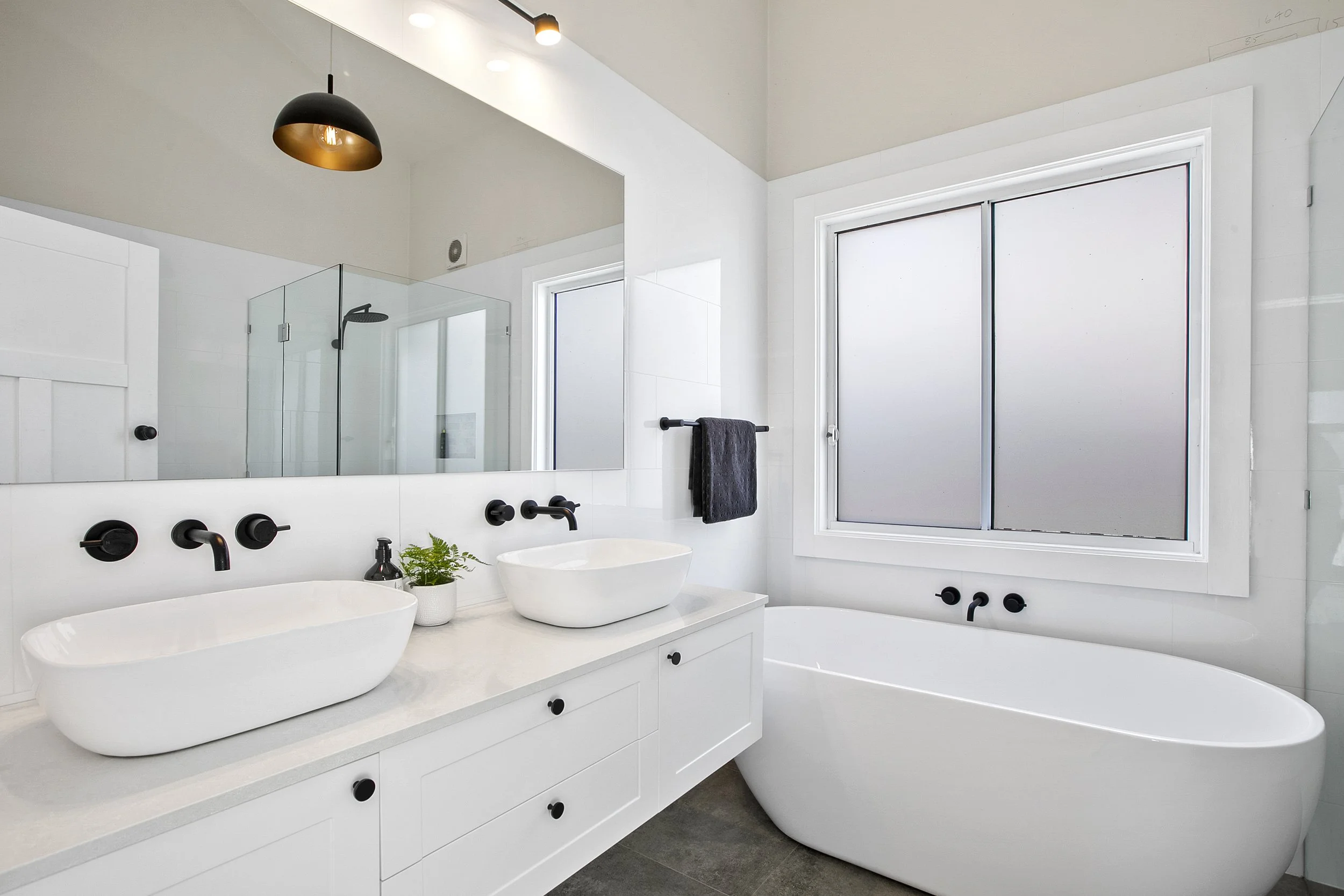 A modern, minimalist bathroom with white walls and double white vessel sinks on a white vanity with black fixtures and knobs. There is a large mirror above the sinks, a small potted plant, and black towels hanging on a towel bar. The bathroom features a large window with sliding frosted glass and a glass-enclosed shower with a black showerhead. A round bathtub is also visible.
