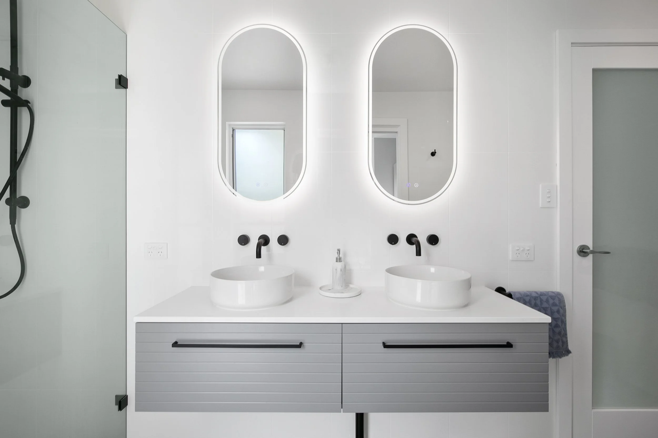Modern white bathroom vanity with two oval mirrors and two vessel sinks, black fixtures, and backlit mirrors, with a window and minimalist decor.