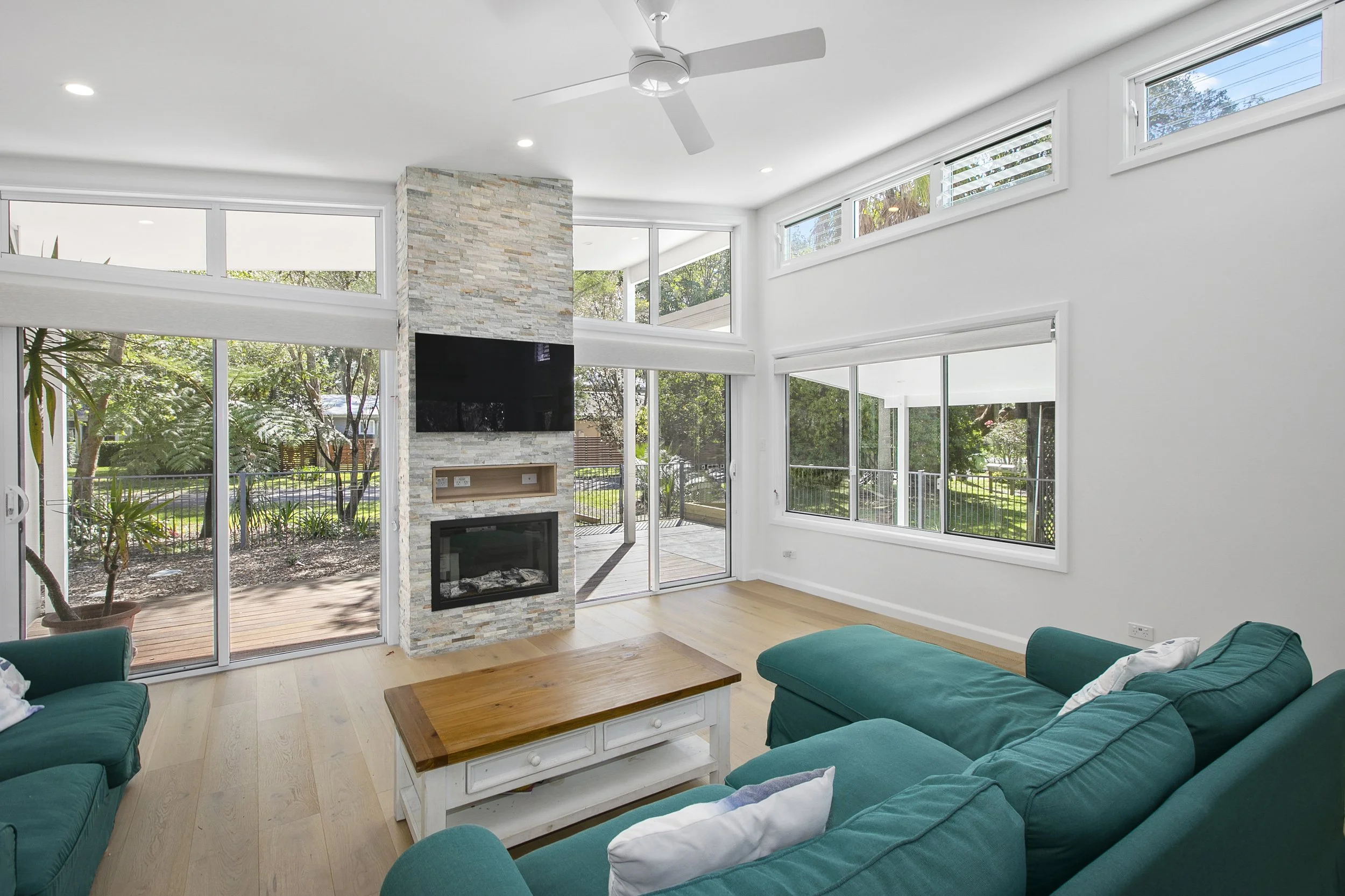 Open-concept living room with large windows, white walls, light wooden flooring, teal sofas, a wooden coffee table, and a stone fireplace with a mounted flat-screen TV. Sliding glass doors lead to an outdoor patio with trees visible outside.