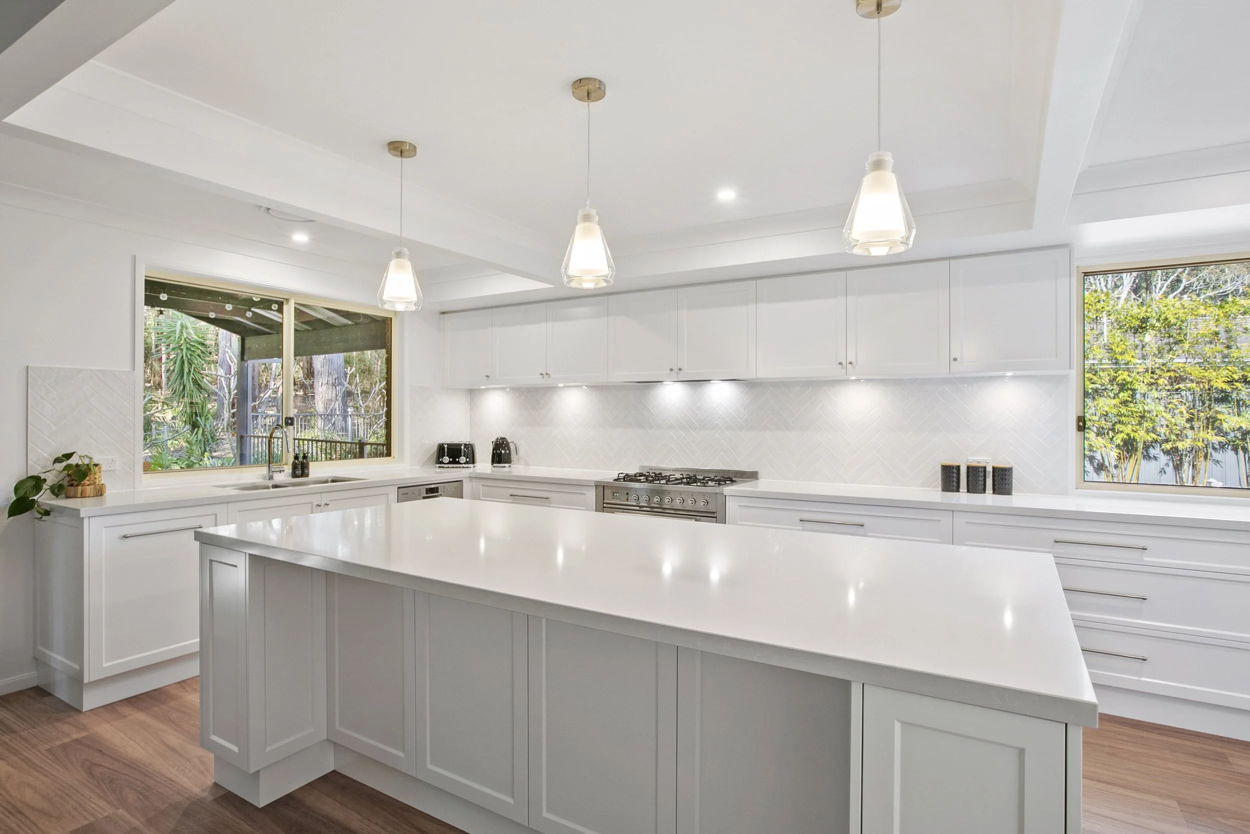 Modern white kitchen with large island, pendant lights, white cabinets, and large windows showing outdoor greenery.
