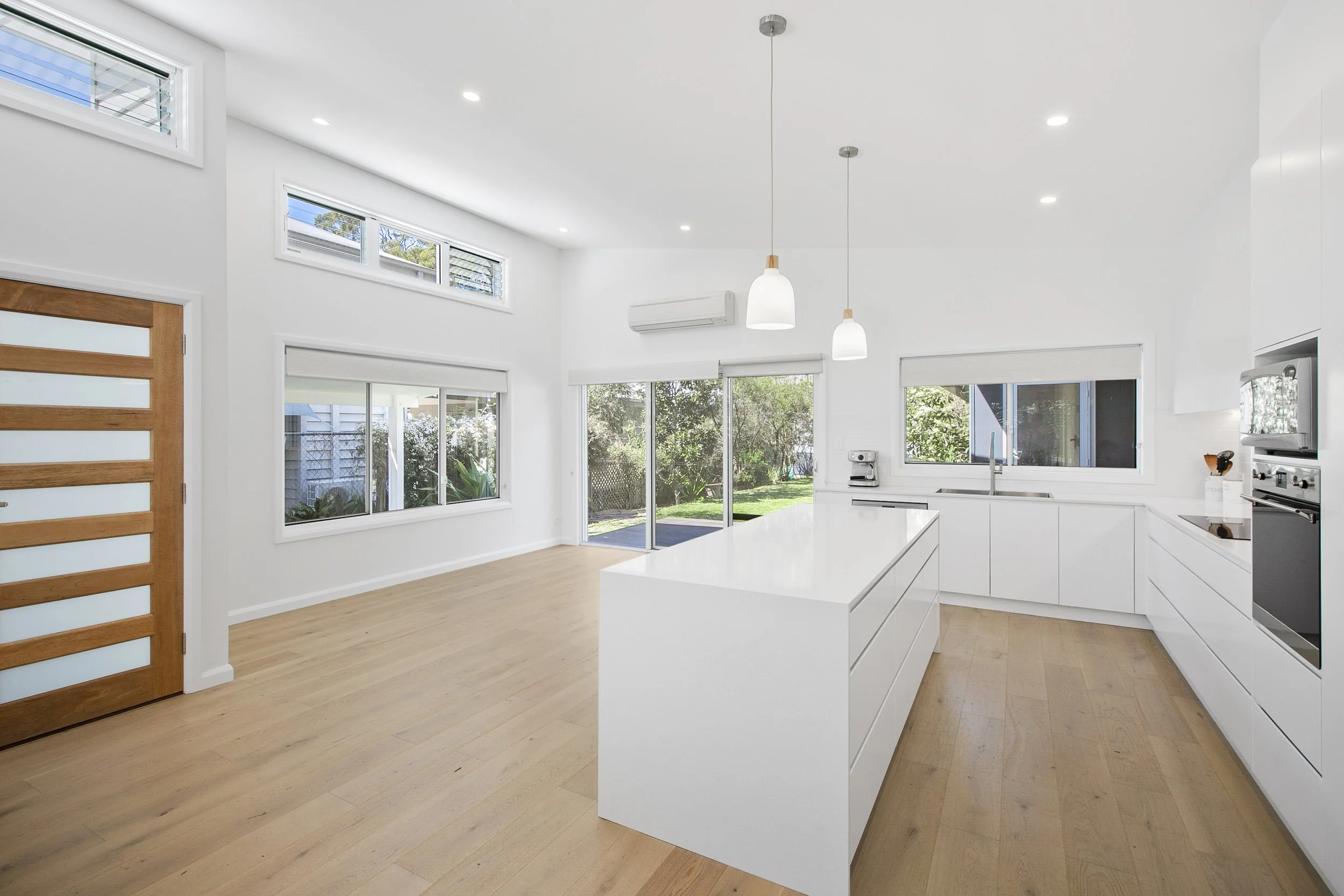 Modern white kitchen with large windows, light wood flooring, and a central island, featuring ceiling lights and sliding glass doors to a backyard.