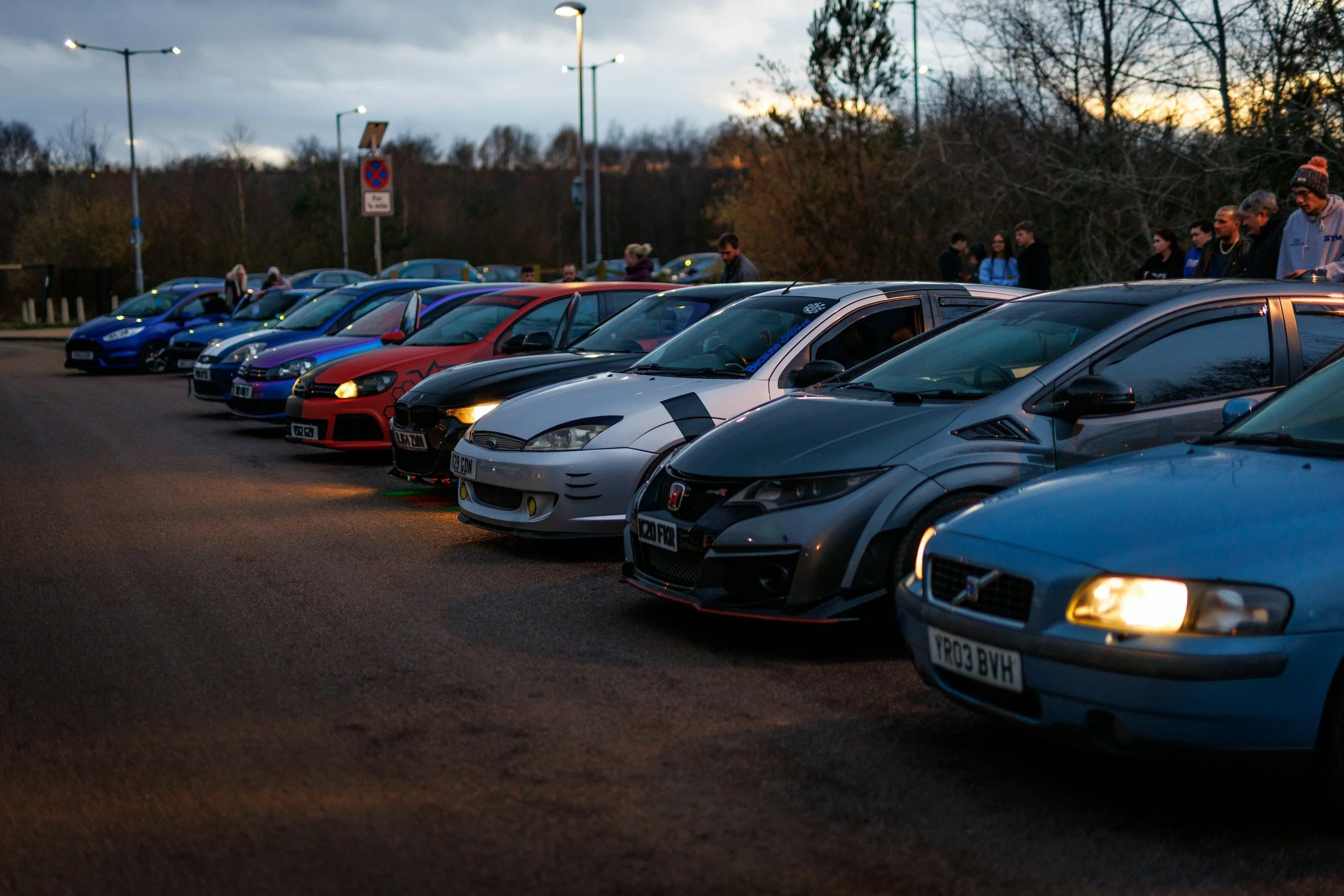 Line of parked cars during dusk with people standing nearby and trees in the background.
