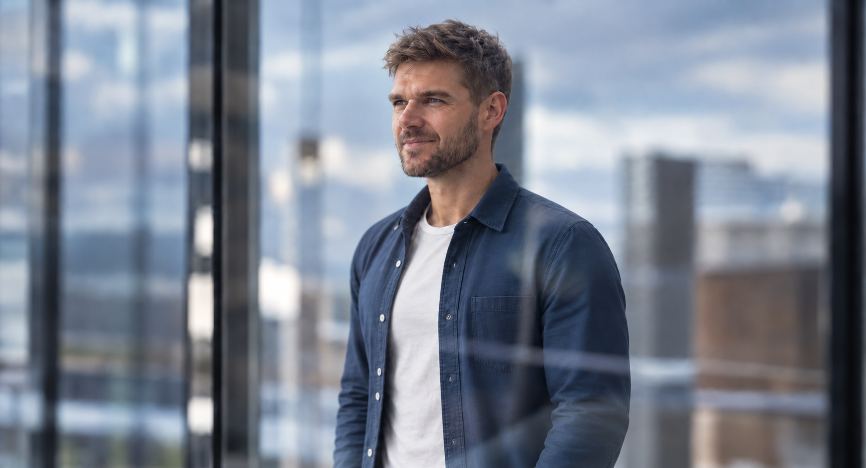 A man with brown curly hair and a beard looking out a window on a cloudy day, wearing a dark blue jacket over a white t-shirt, with city buildings in the background.