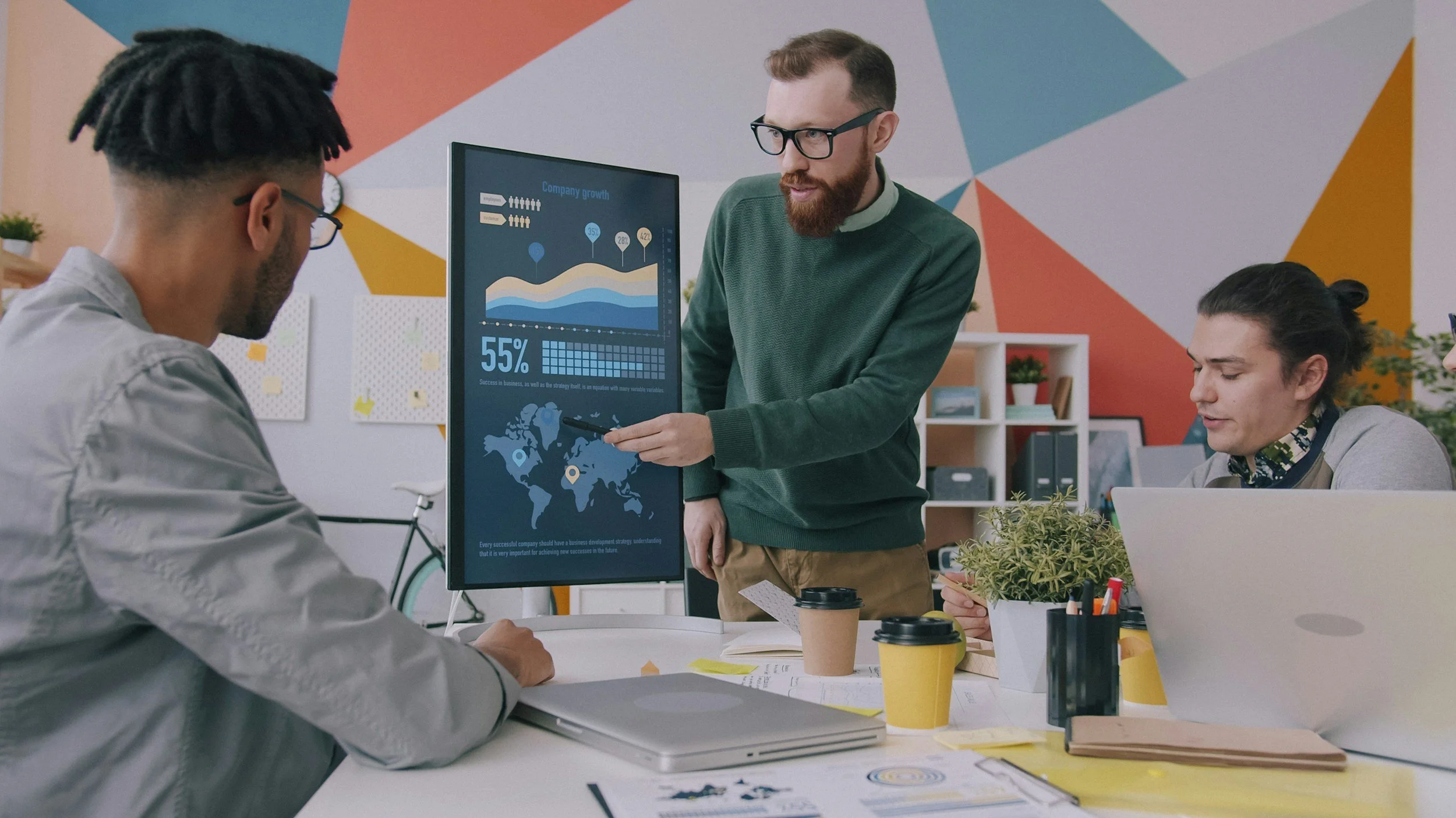 A business meeting in a modern office with three people. One person is presenting data on a large screen, showing growth charts, a map, and a percentage. The other two are seated, with laptops and documents. The background features colorful geometric wall art and shelves with plants and decor.