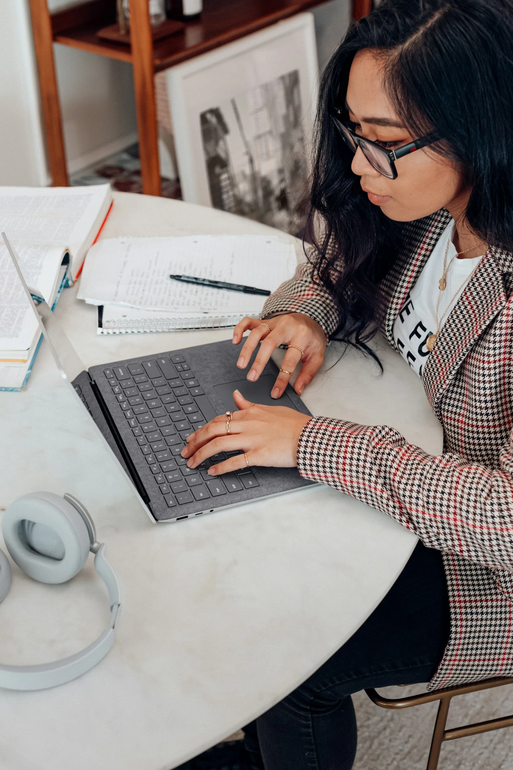 A woman with long black hair, glasses, and rings on her fingers is working at a desk. She is using a laptop with a black keyboard. On the desk, there are open books, a spiral notebook, and a pen. A pair of white headphones is placed on the desk near the laptop. In the background, there is a wooden shelf with various items and a black and white photo leaning against the wall.