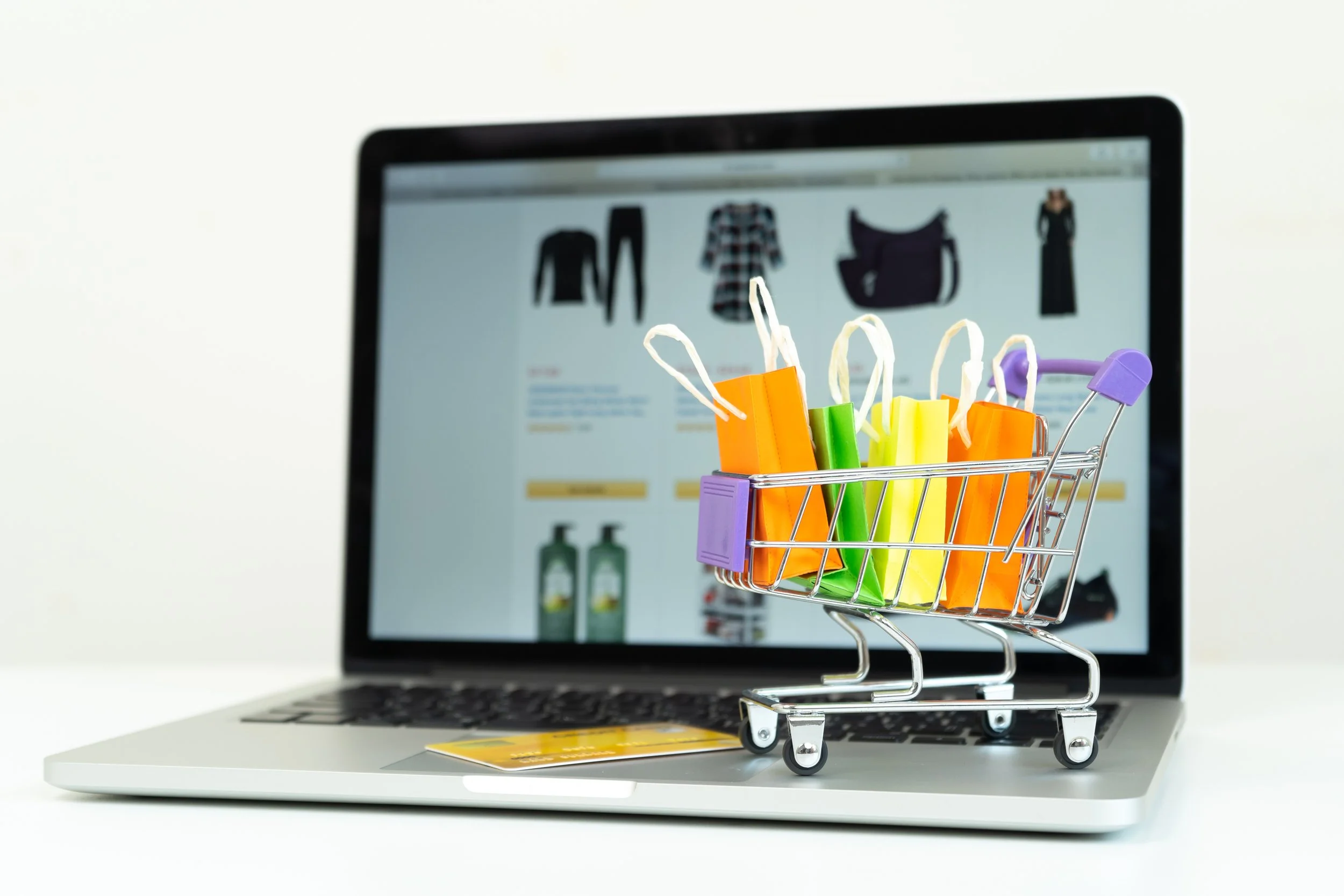 Miniature shopping cart filled with colorful shopping bags sitting on a laptop keyboard, with a computer screen displaying clothing items in the background.