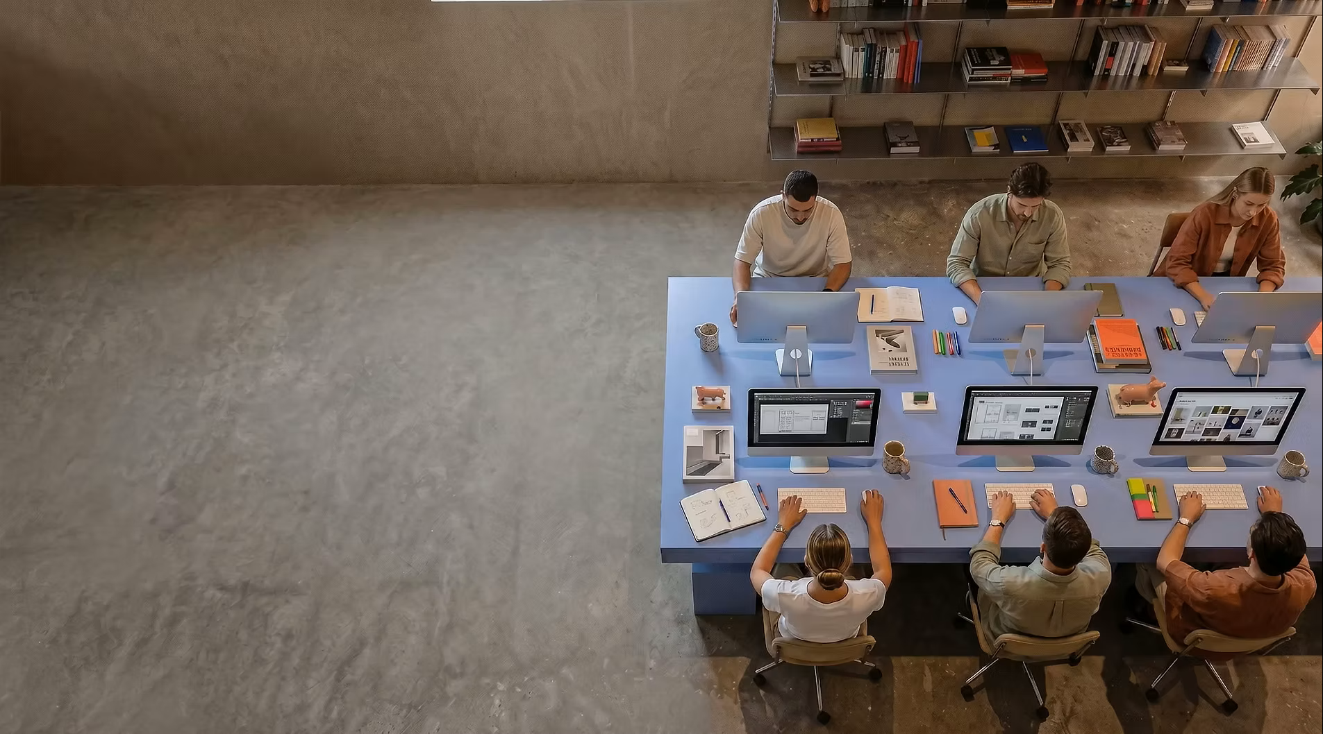 Top-down view of a group of people working at a large blue desk with multiple computers and various office supplies.