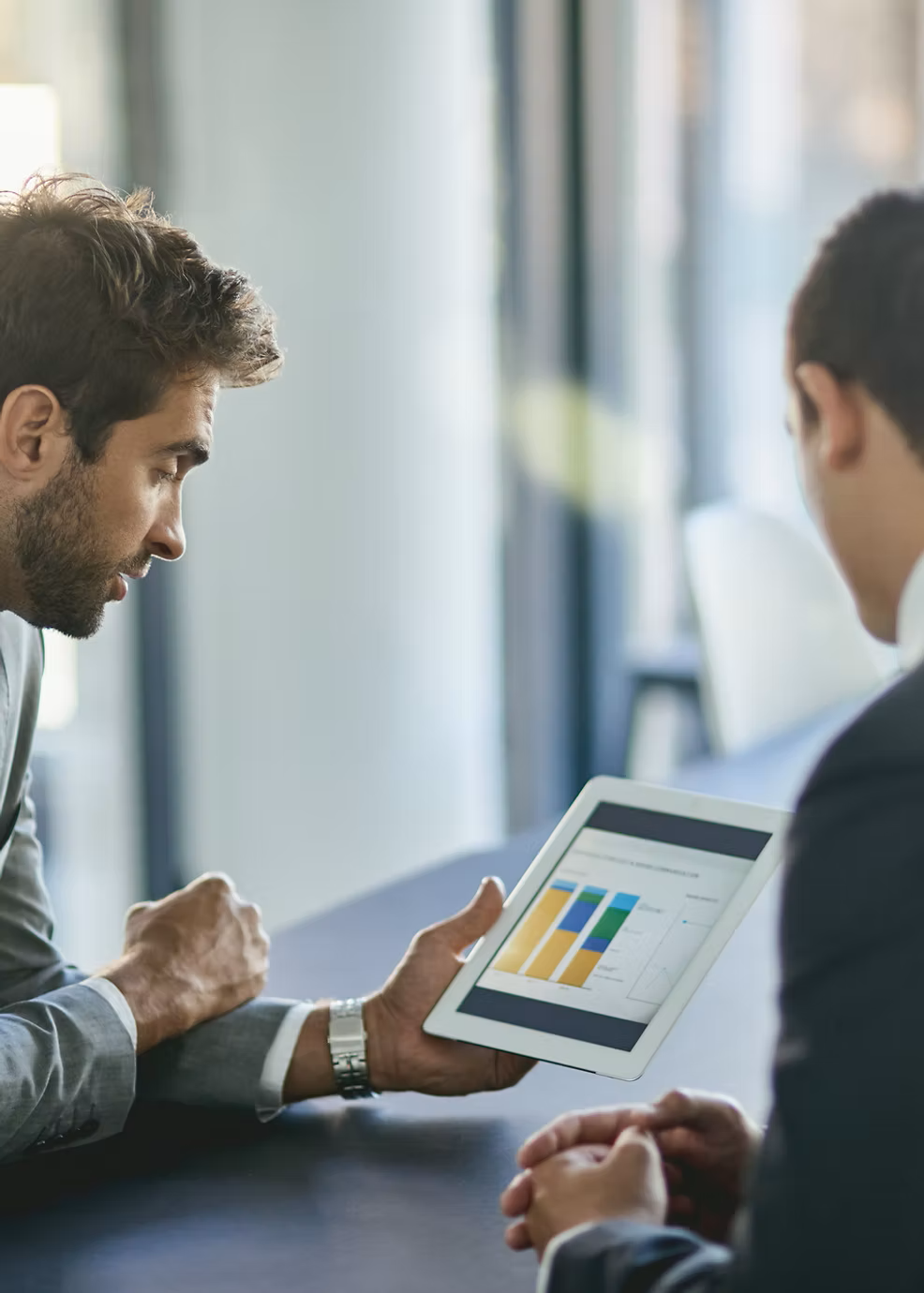 Two businessmen having a discussion, with one showing data charts on a tablet, in a modern office setting.