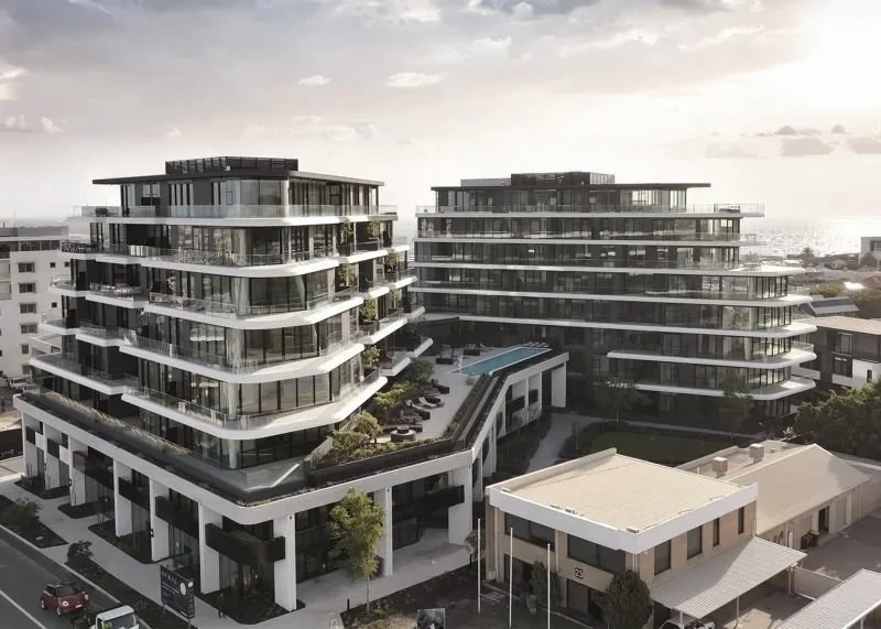 Two modern, multi-story residential buildings with glass balconies and a shared rooftop area, pool, and outdoor seating, set in an urban area near the coast.