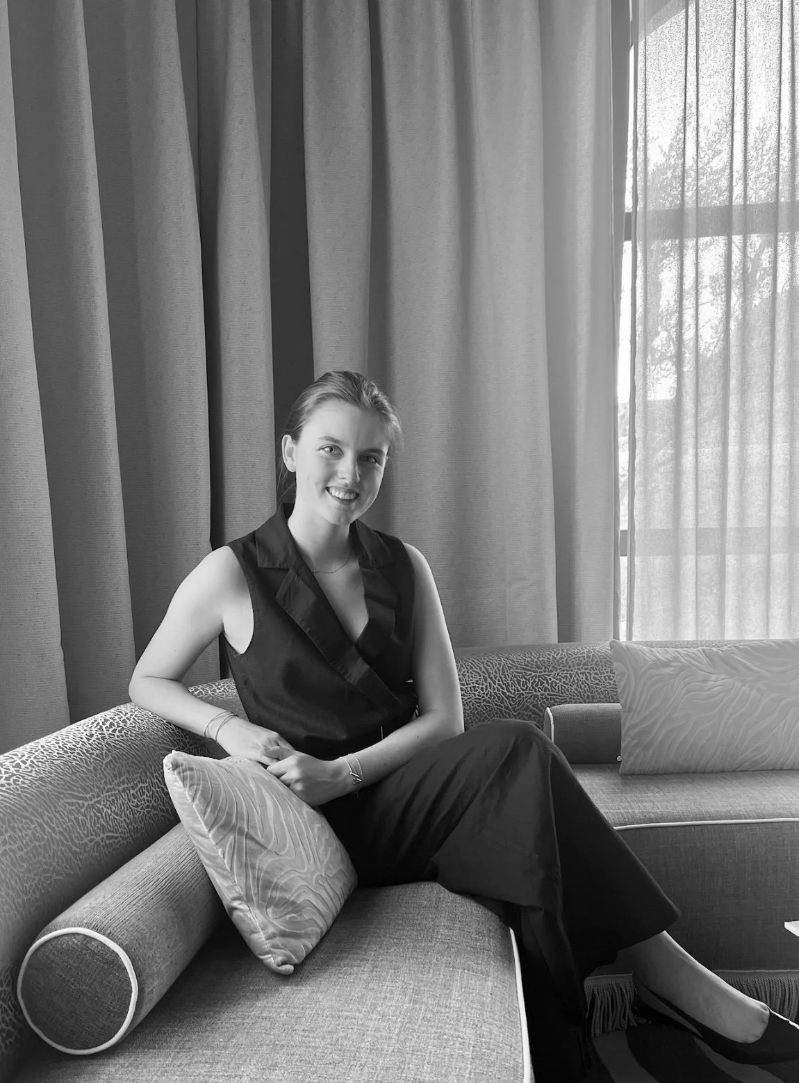A woman sitting on a sofa in a room with curtains and a window, smiling at the camera, in black and white.
