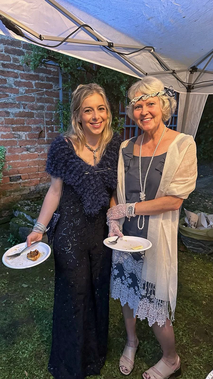 Two women in elegant dresses holding food plates at an outdoor event.