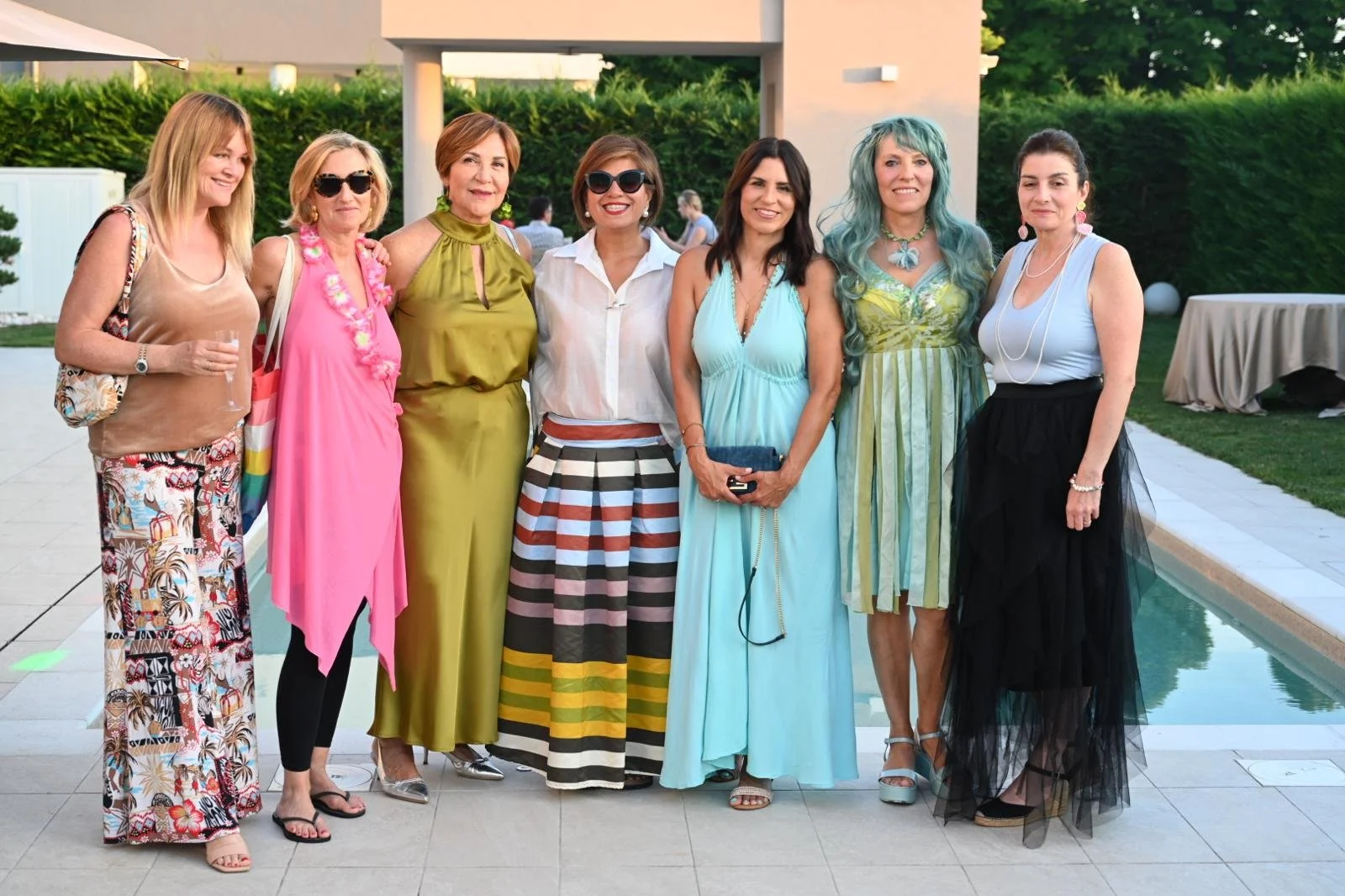 Group of eight women dressed in colorful summer outfits standing together by a pool at an outdoor party or gathering.