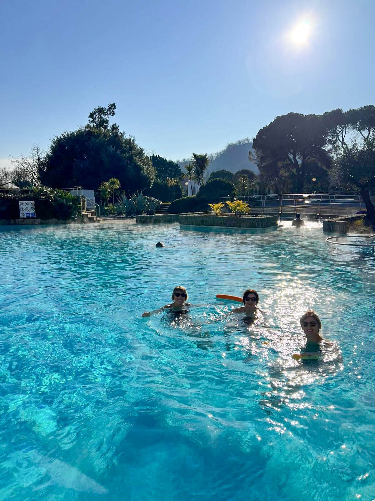 People swimming and enjoying a hot spring pool outdoors on a sunny day with trees and hills in the background.