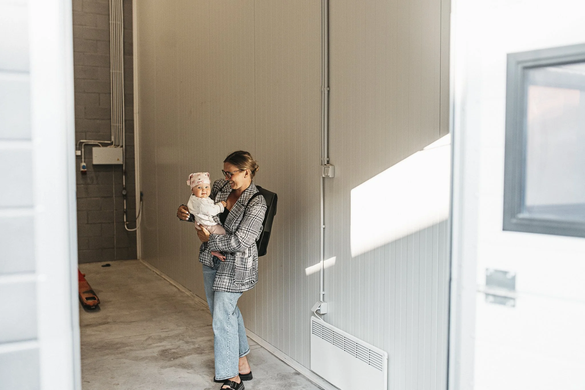 A woman with glasses and a checked coat holding a baby with a pink hat, standing inside a building with beige walls and a vent, smiling at each other.