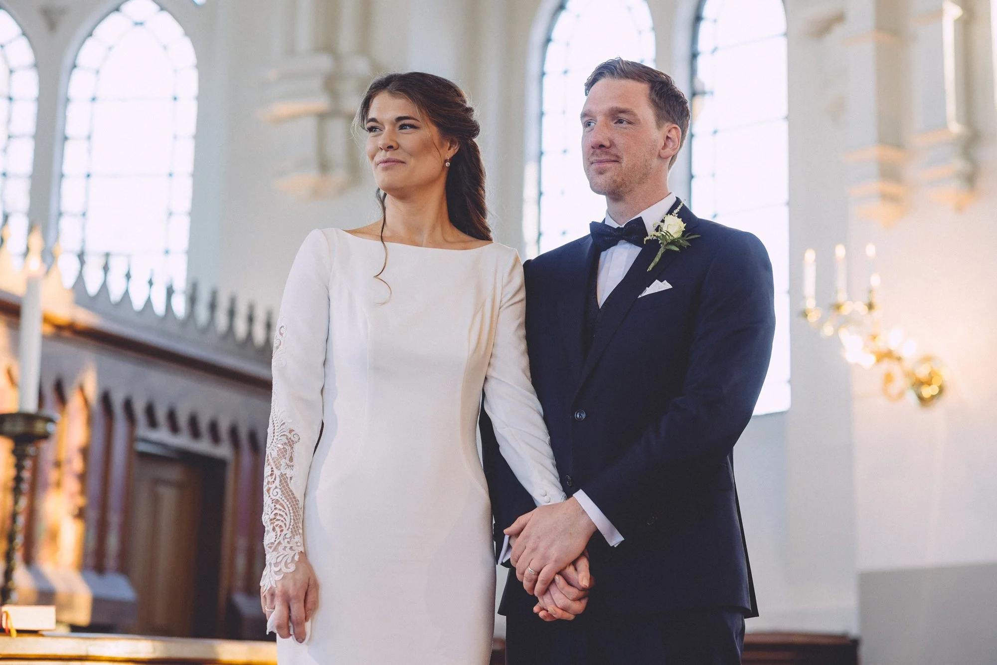 En nygift kvinna och man står tillsammans i en kyrka under deras bröllopsceremoni. Kvinnan bär en vit brudklänning med utsmyckade ärmar och mannen bär en svart tuxedo med svart fluga.