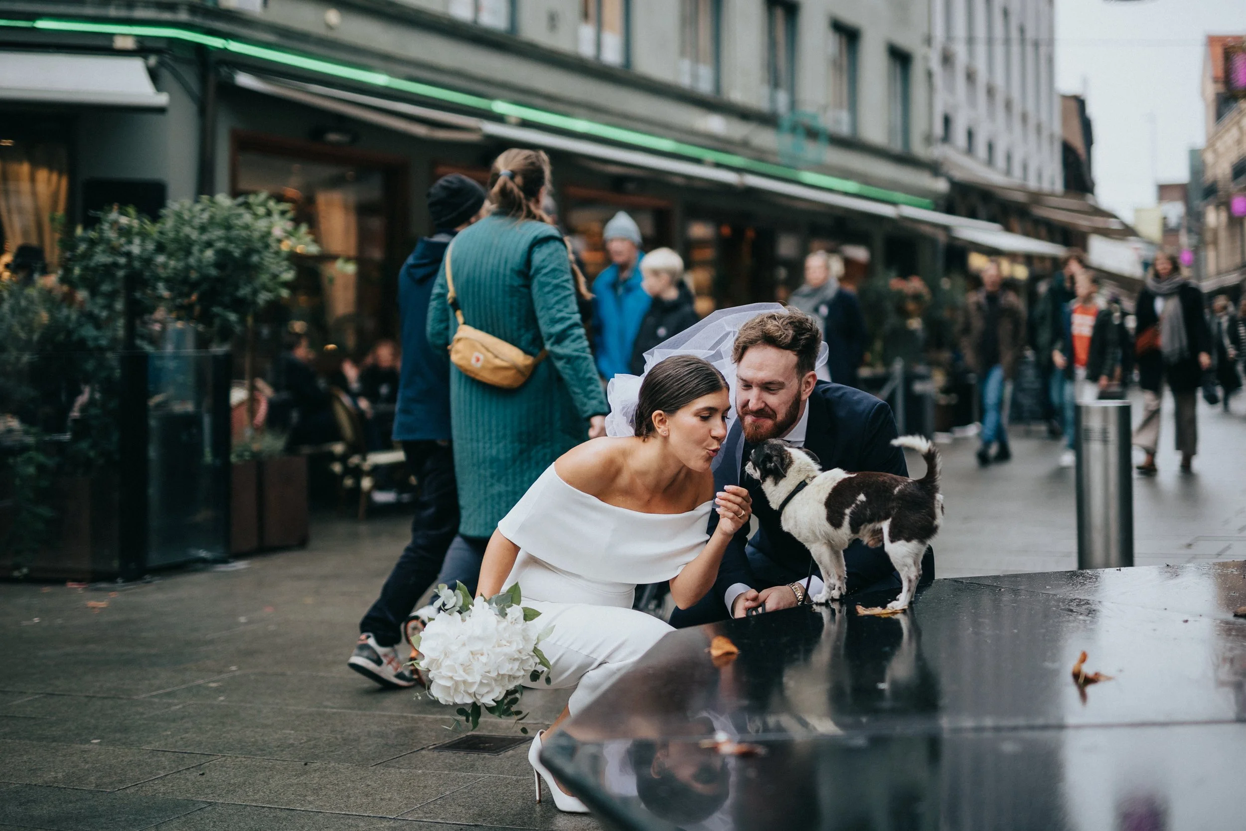 Nygifta brud och brudgum sitter på en stadspromenad och leker med en hund, omgiven av människor och stadsmiljöer.