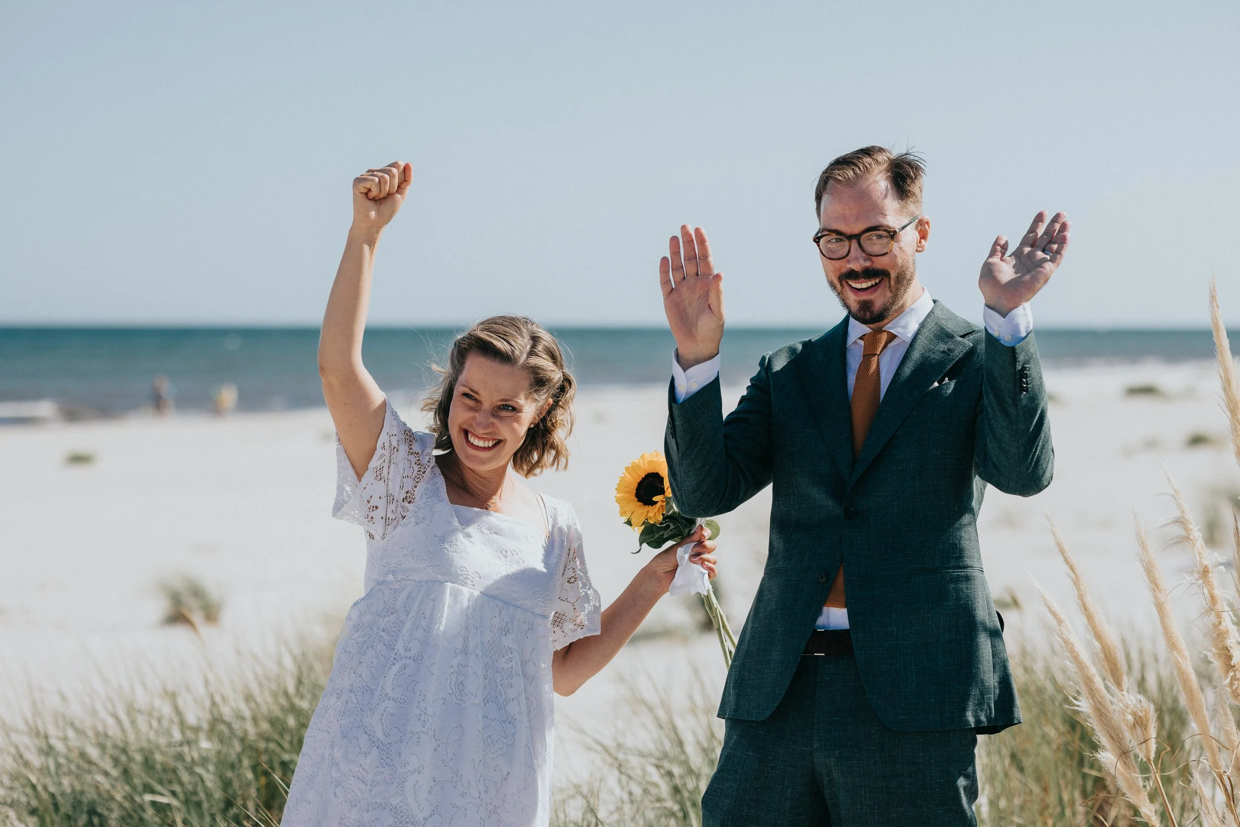 En kvinna i vit klänning och en man i kostym på stranden, båda ler och partar, kvinna håller en solros och mannen viftar med händerna.