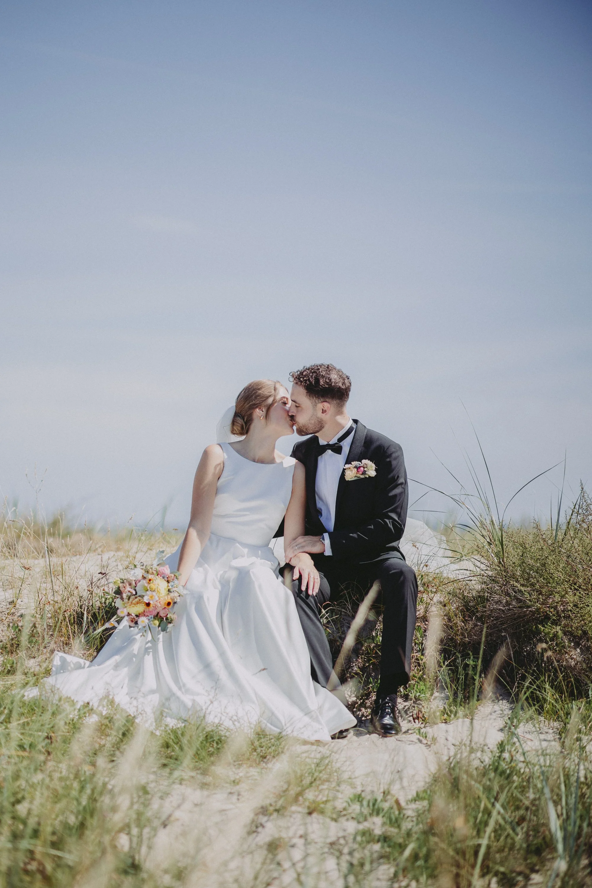 En brud och en brudgva kyssas på en strand, omgiven av gräs och blå himmel.