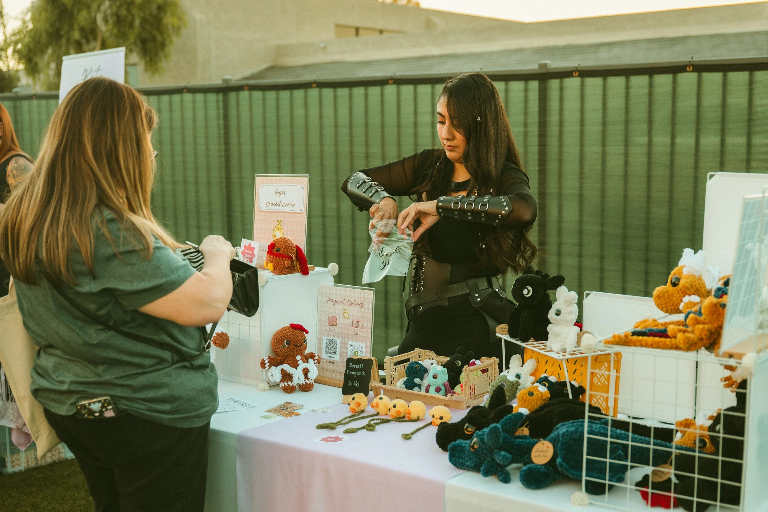 A woman with long dark hair wearing a black outfit with studded sleeves is looking at handmade stuffed animal toys at a craft fair booth. The booth has a table with various crocheted animals and a sign that says "Gigap Crochet Corner". A customer with long blonde hair wearing a green shirt is browsing the display.