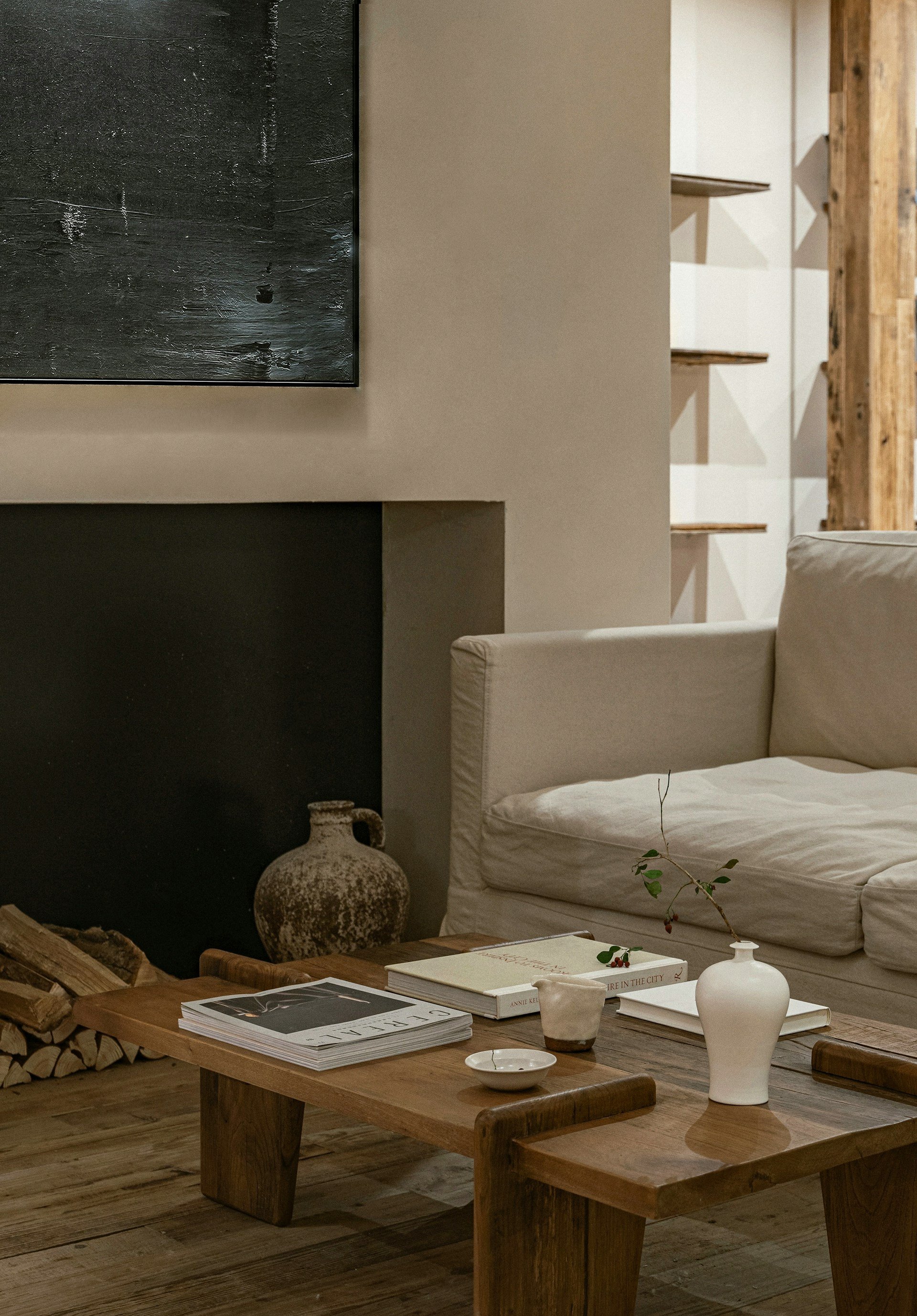 Premium Squarespace Template for Interior Designers. Living room with a beige couch, a wooden coffee table with books, a white vase with a branch, a small white dish, a cup, and a large vase near a black fireplace with stacked logs.
