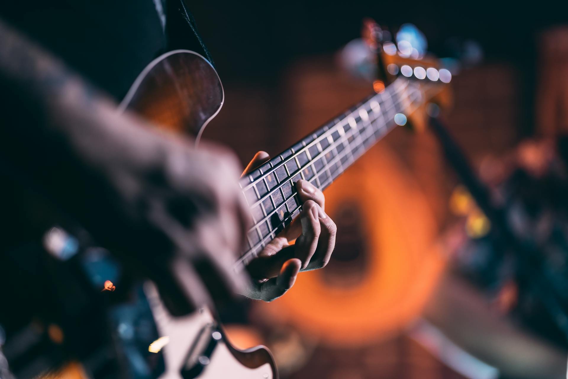 A person playing an acoustic guitar, with focus on their hand on the fretboard, in a dimly lit setting with blurred colorful lights in the background.