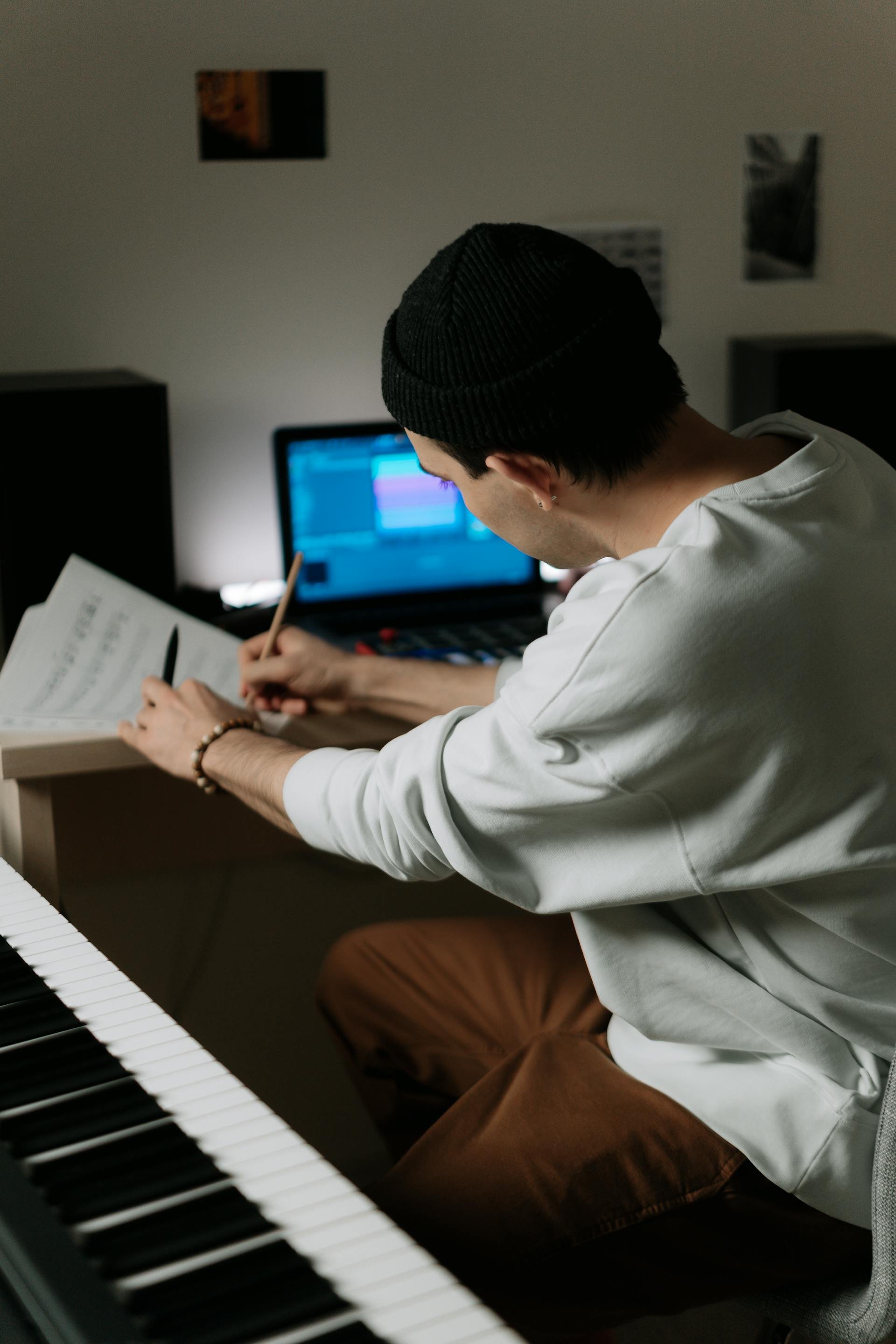 Young man playing piano, wearing a black beanie and white shirt, sitting at a digital keyboard in a room with wall art.